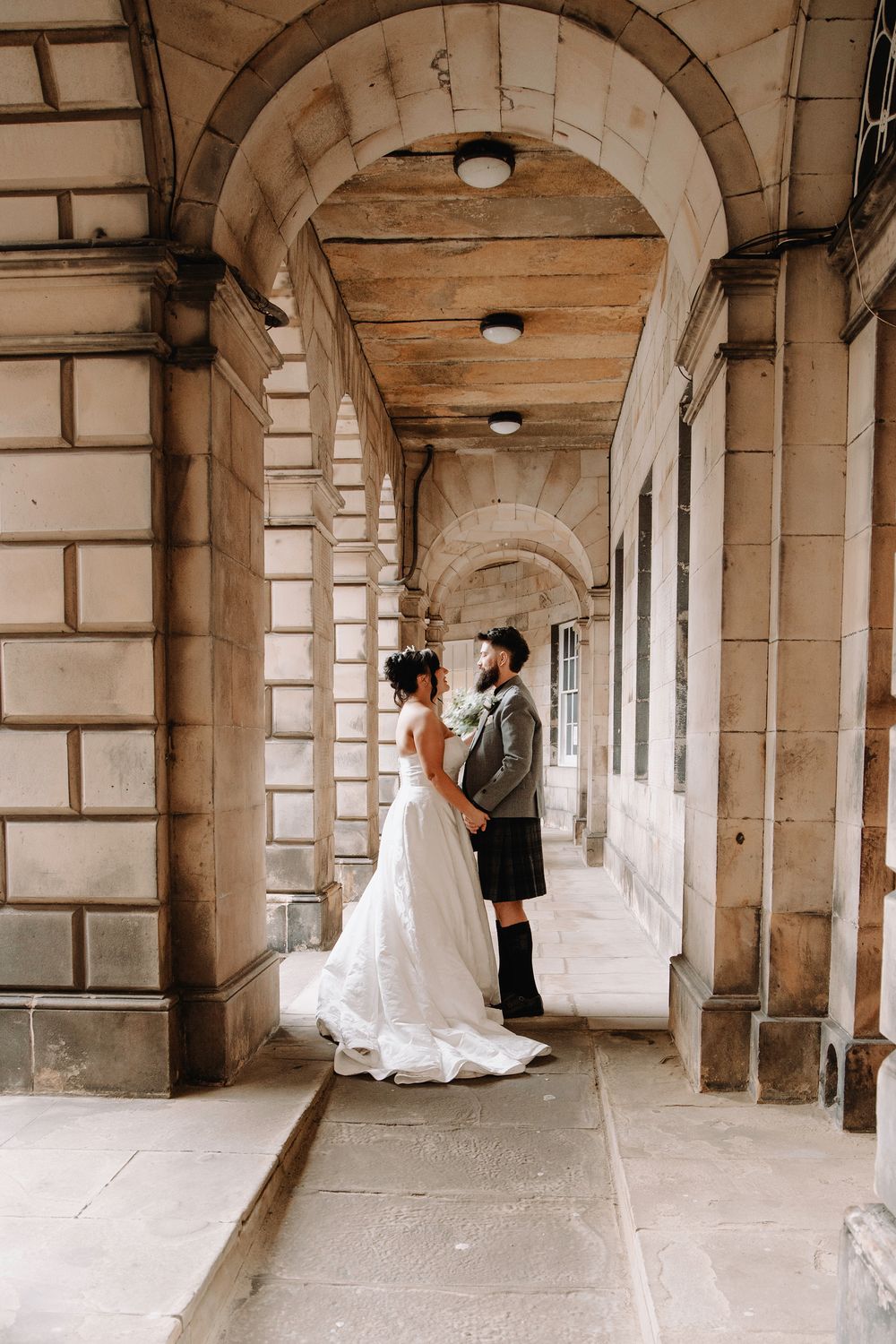 Bride and groom stand facing each other under stone arches of a historic building, bride in white gown, groom in kilt.
