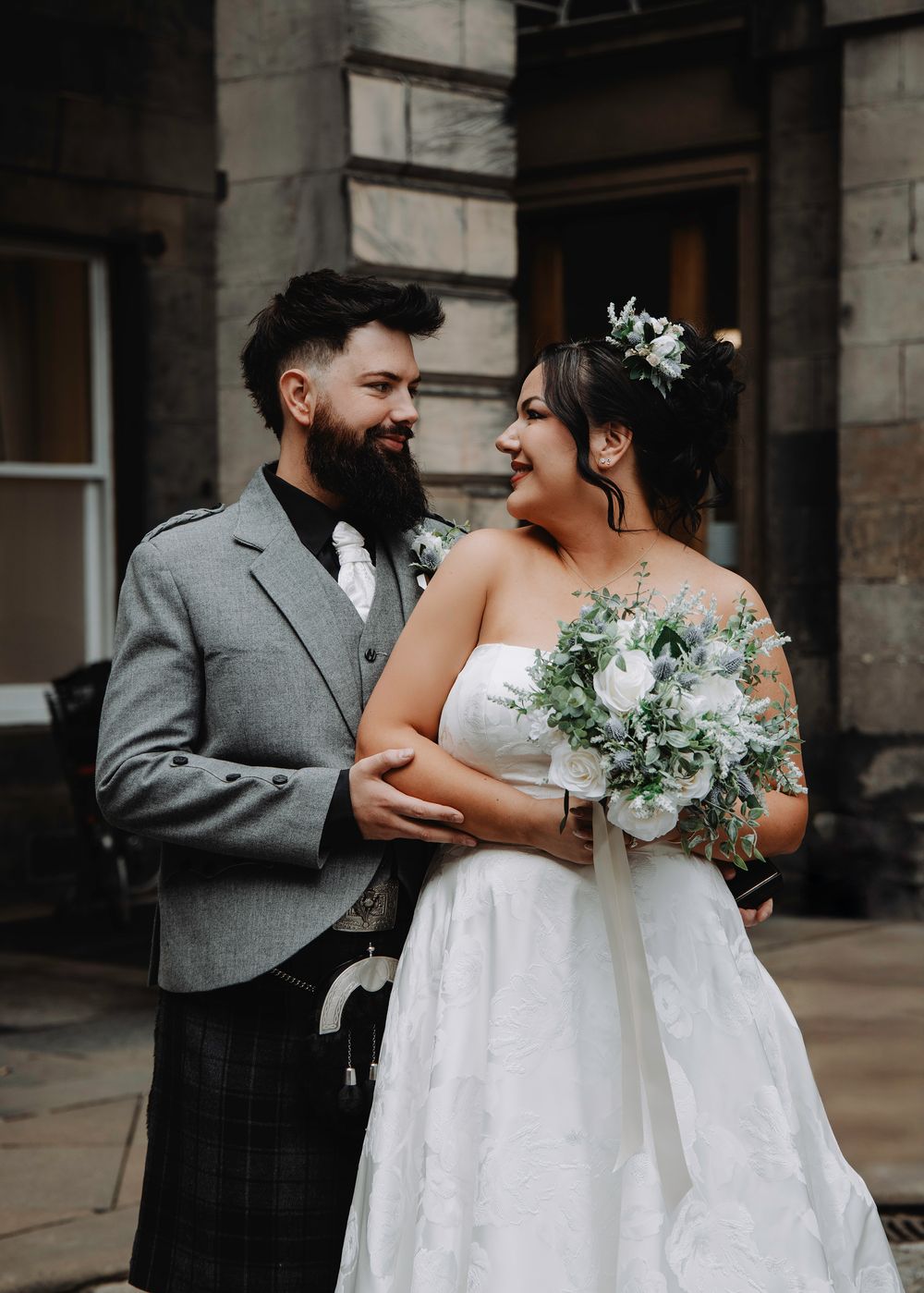 Bride and groom sharing a loving glance, groom in Scottish kilt, bride holding white floral bouquet outside stone building.