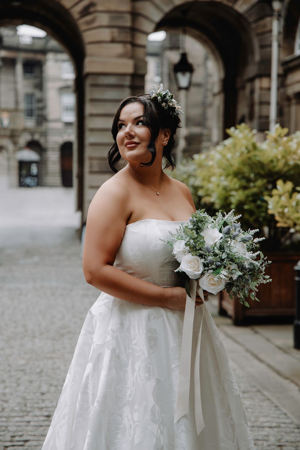 Elegant bride in strapless white gown holding white and green bouquet, posing under stone archway in historic city setting.
