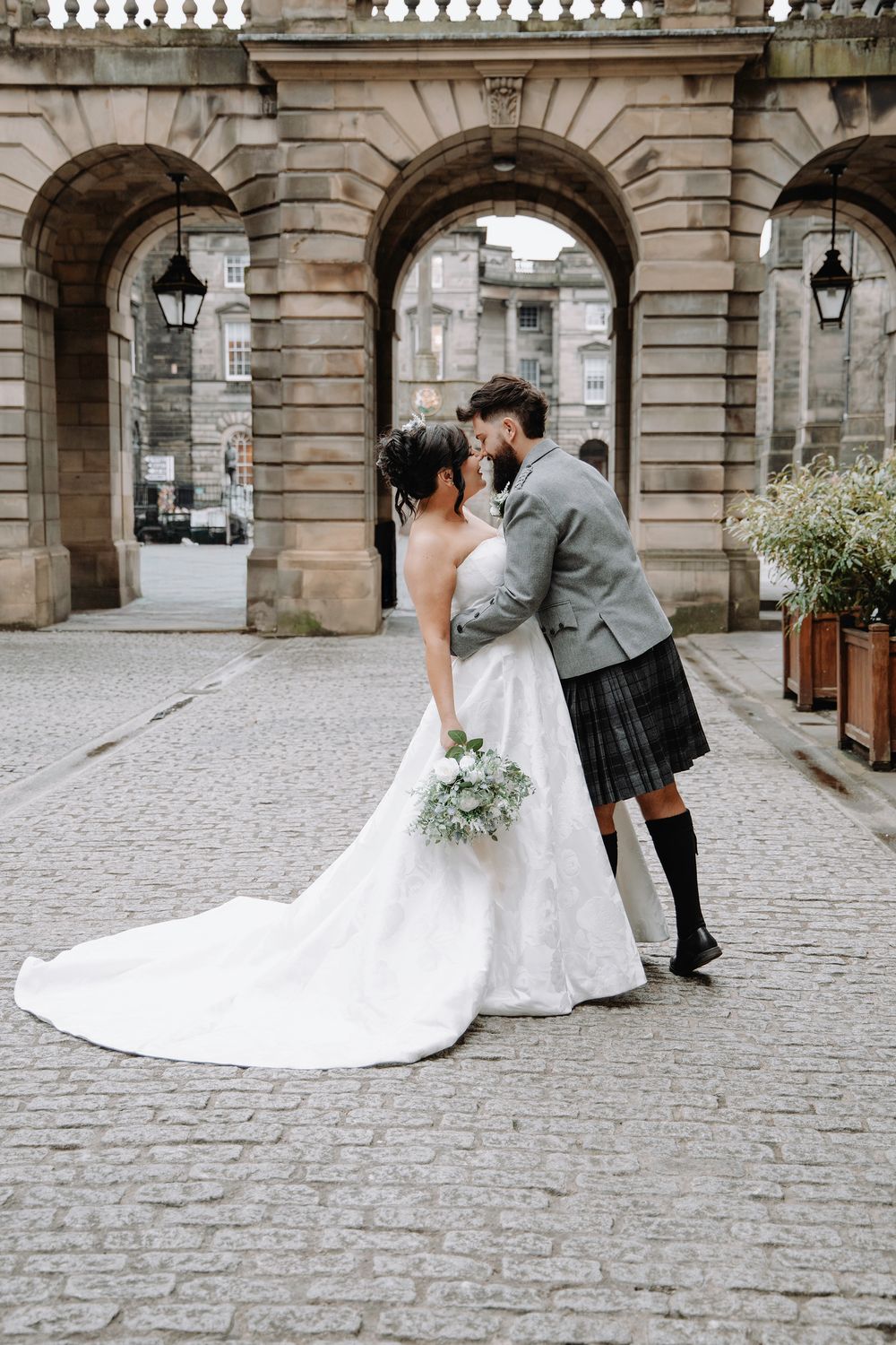Bride and groom share a kiss on cobblestone street, bride in white lace gown with train, groom in grey jacket and tartan kilt.