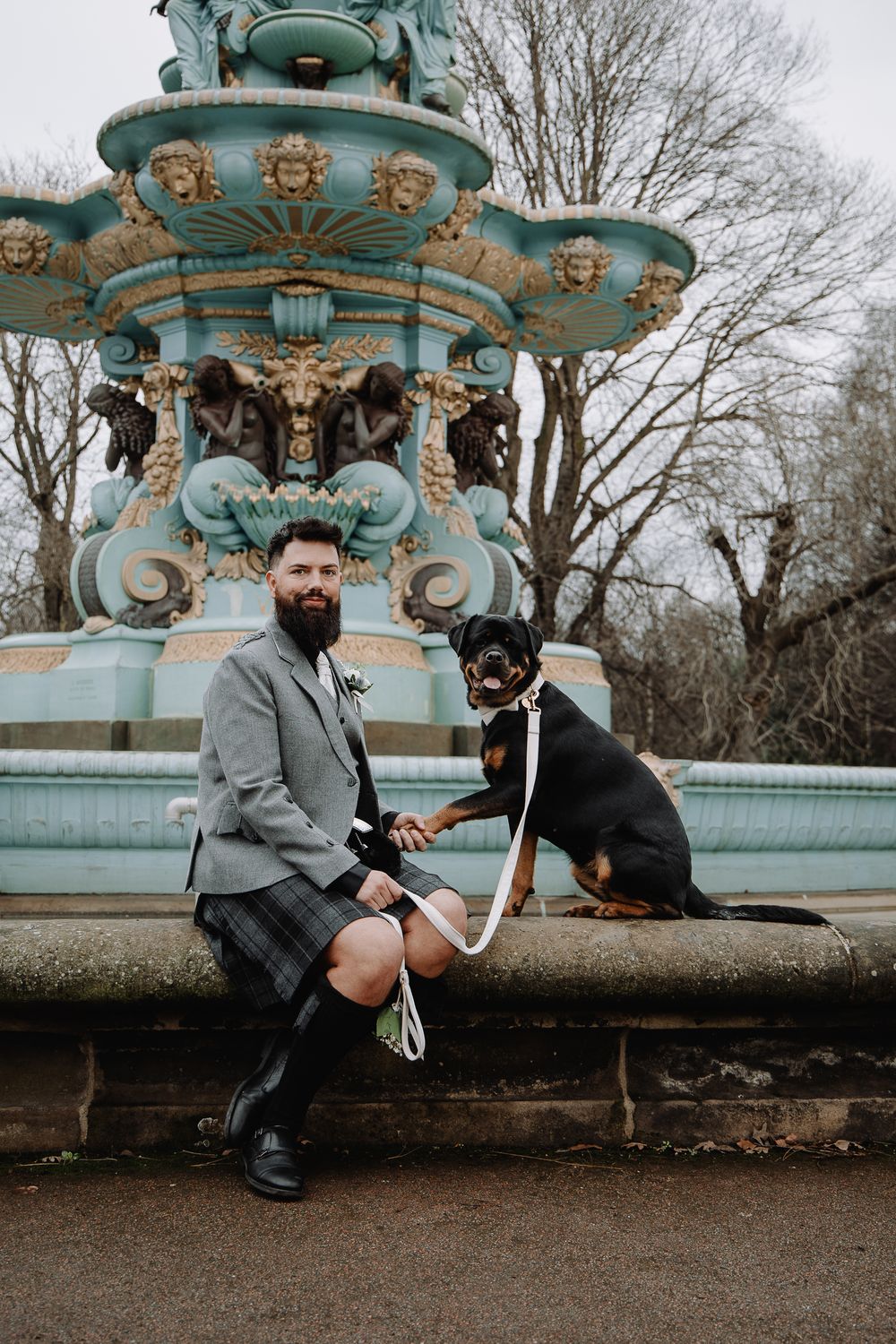 Bearded groom in kilt sits with Rottweiler by ornate teal fountain in Edinburgh park.