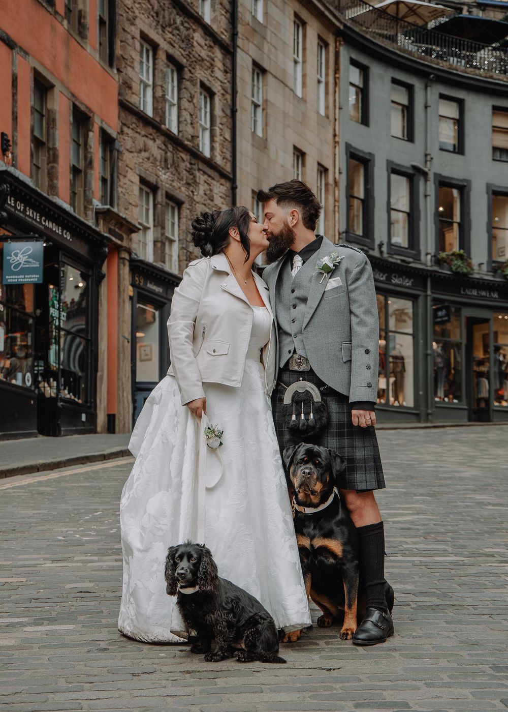 Newlyweds kissing on cobblestone street in Edinburgh, accompanied by two dogs, groom in traditional Scottish kilt attire.