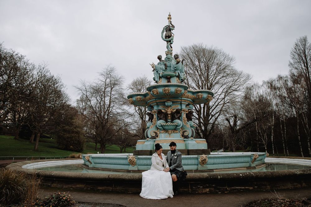 Bride and groom sitting together by a ornate teal fountain in a park on their wedding day.