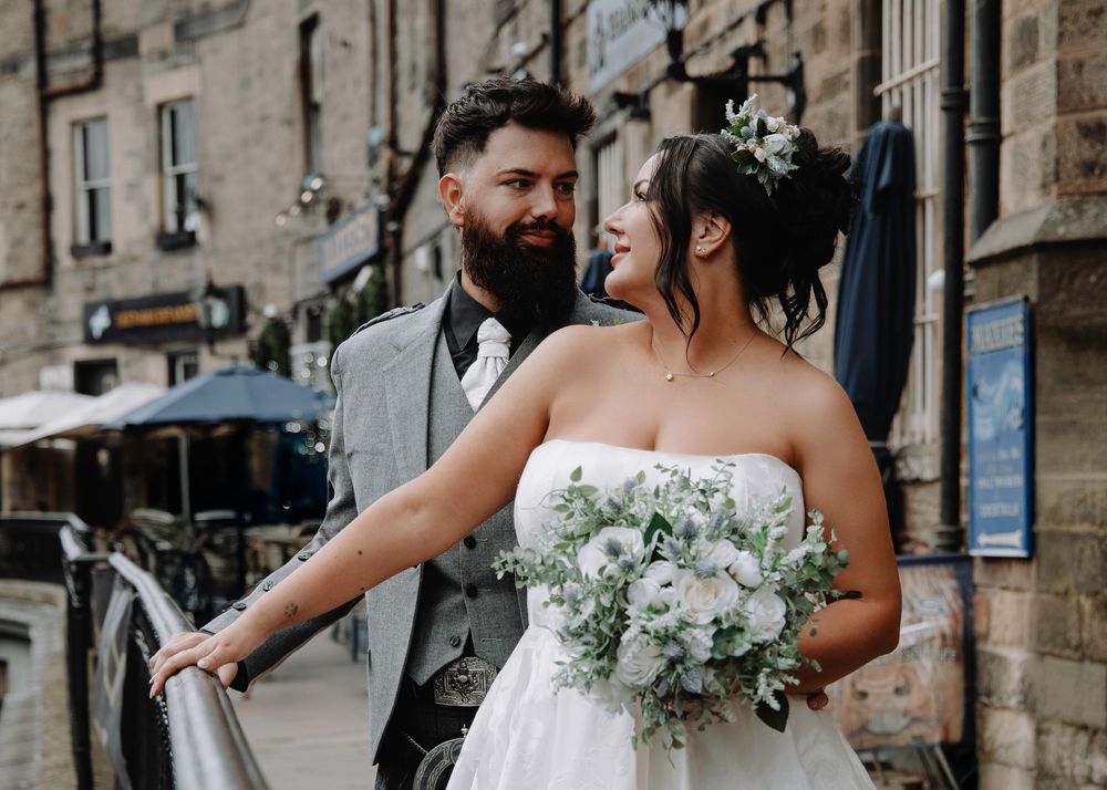 A bride and groom pose outdoors on their wedding day, she holds a white bouquet, wearing a strapless gown.