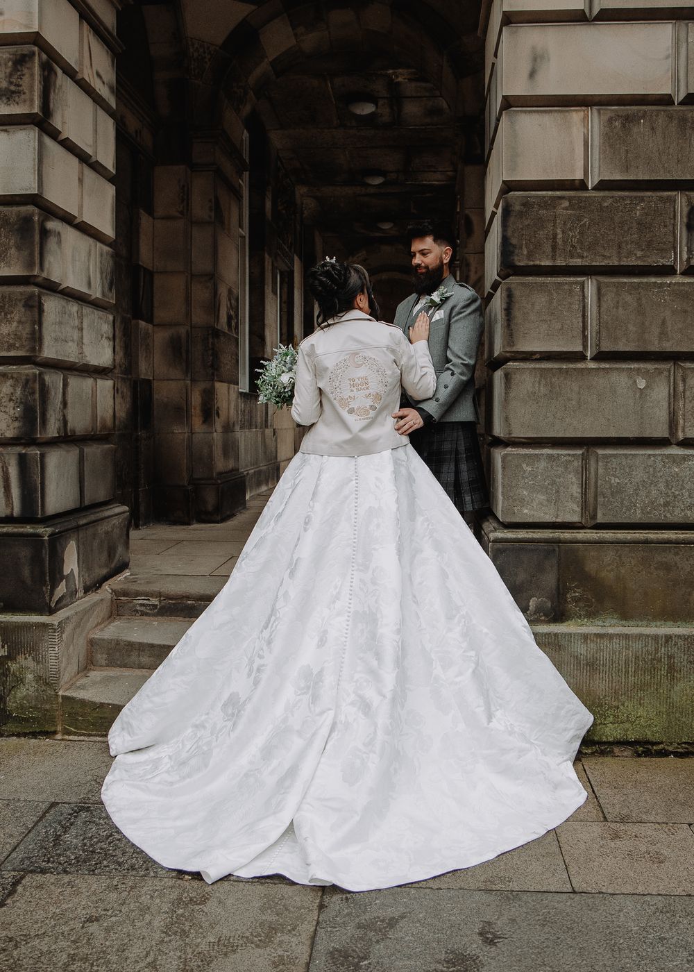 Bride in stunning white ball gown with embroidered jacket and long train, standing with groom at historic stone building.