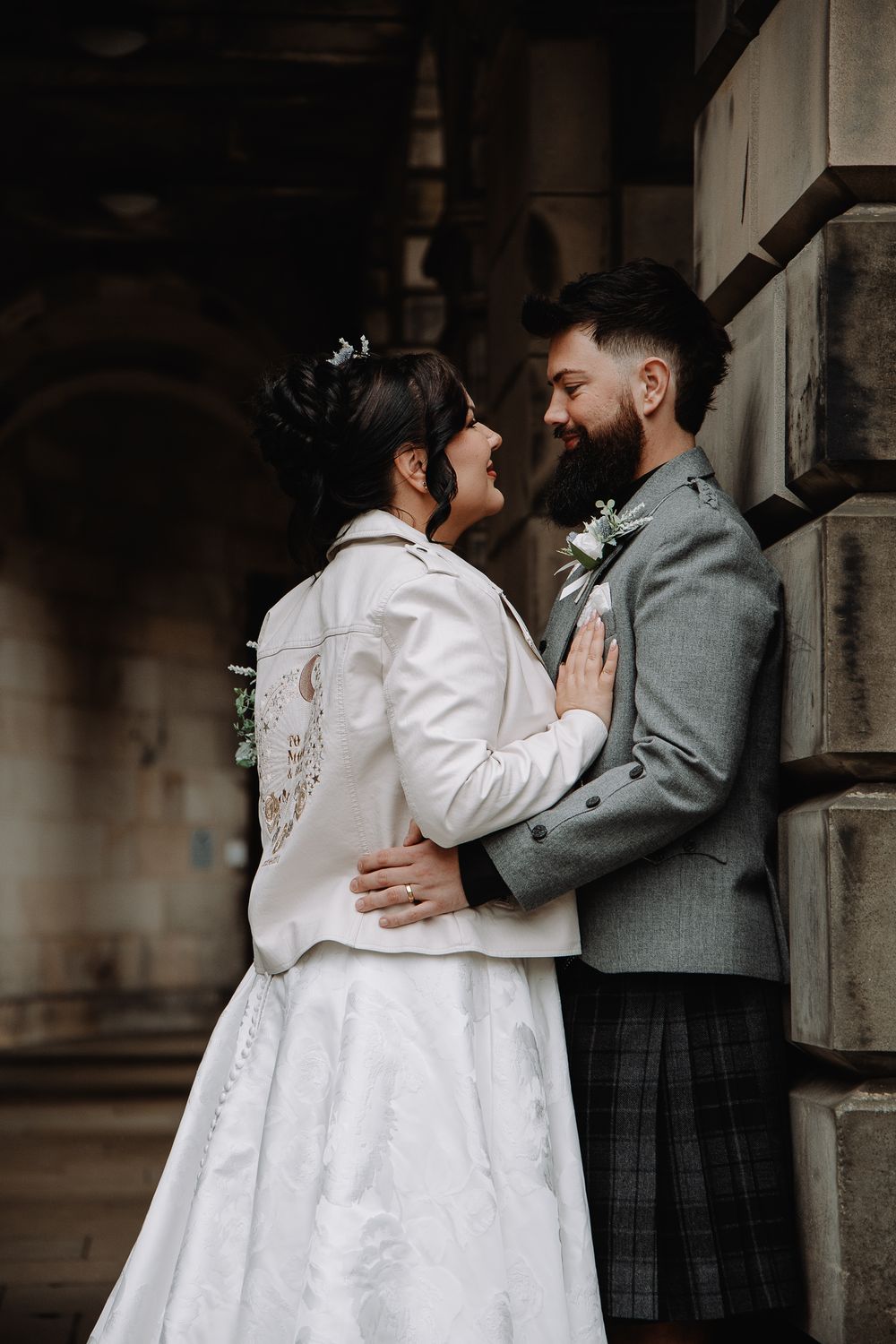 Smiling bride in white lace gown and jacket embraces bearded groom in grey jacket and tartan kilt against stone pillar.