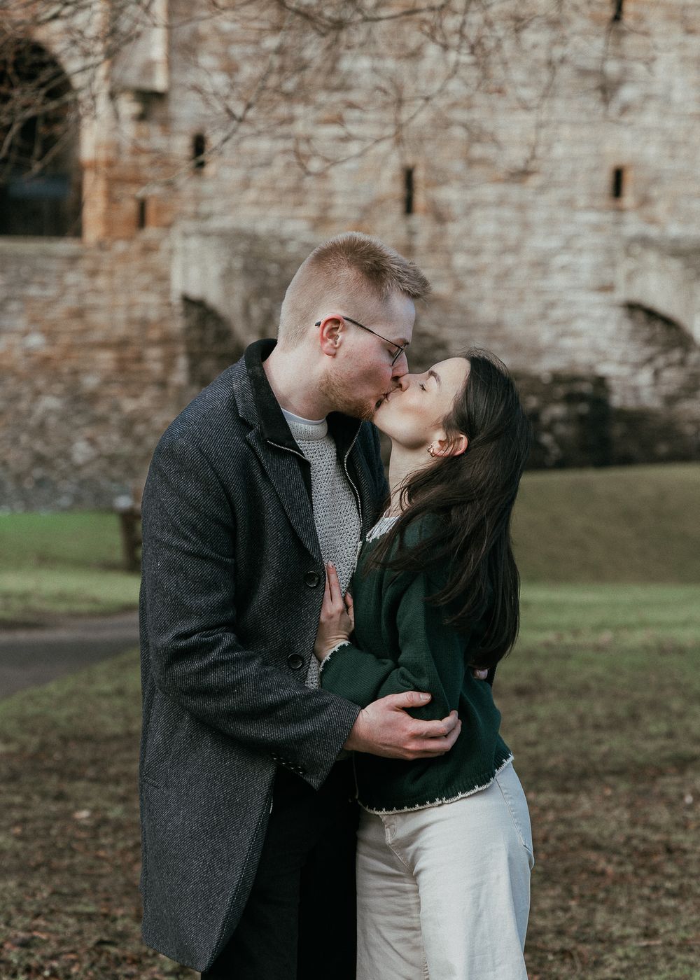 Couple sharing a romantic kiss outdoors near a historic stone castle in winter.