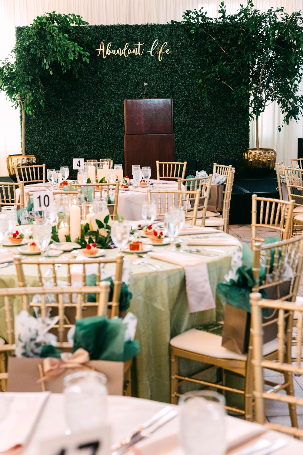 Elegant wedding reception tables set with gold chairs and floral centerpieces against a lush green hedge backdrop.