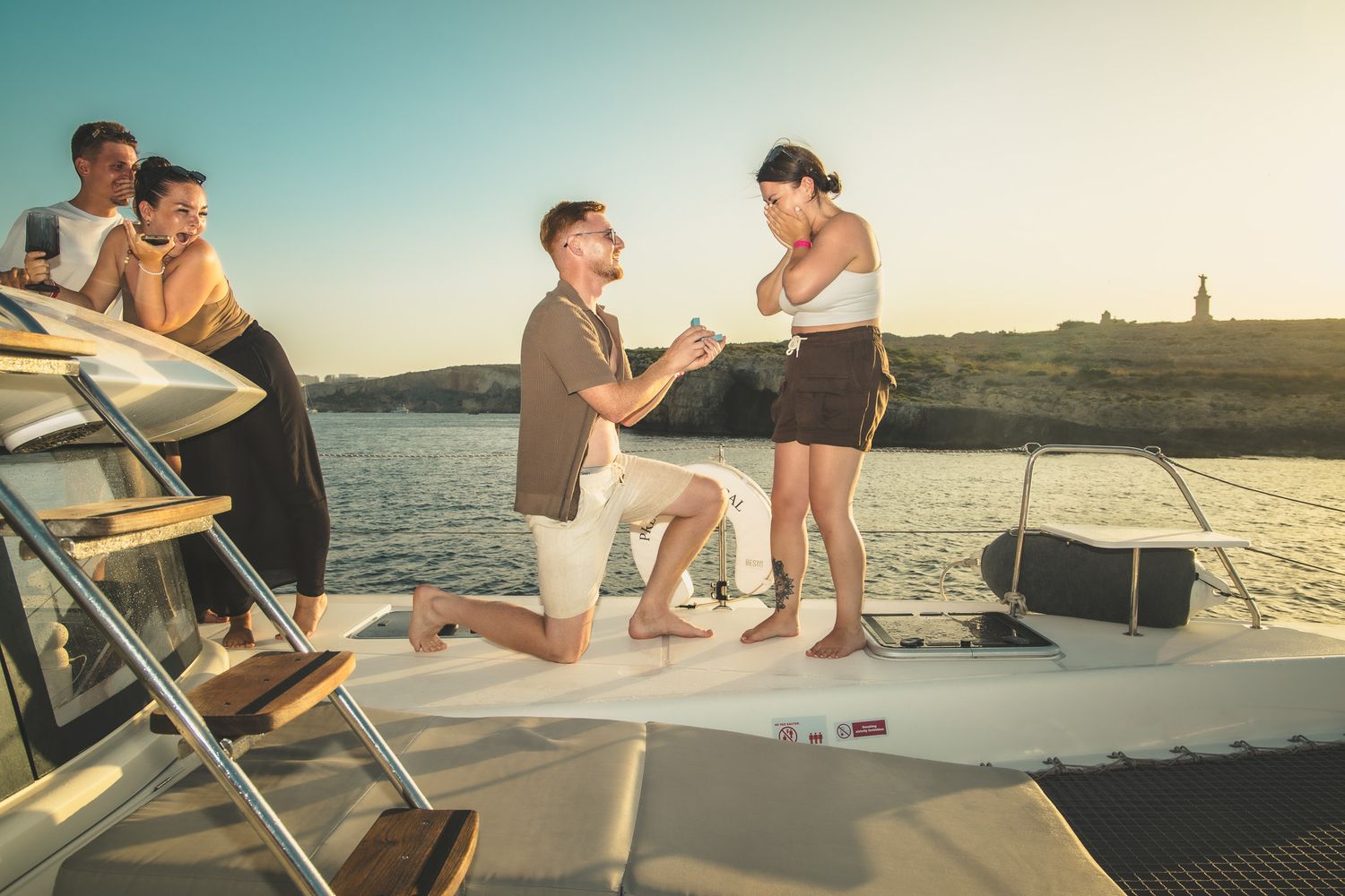 Man proposes on a yacht at sunset while friends watch nearby.