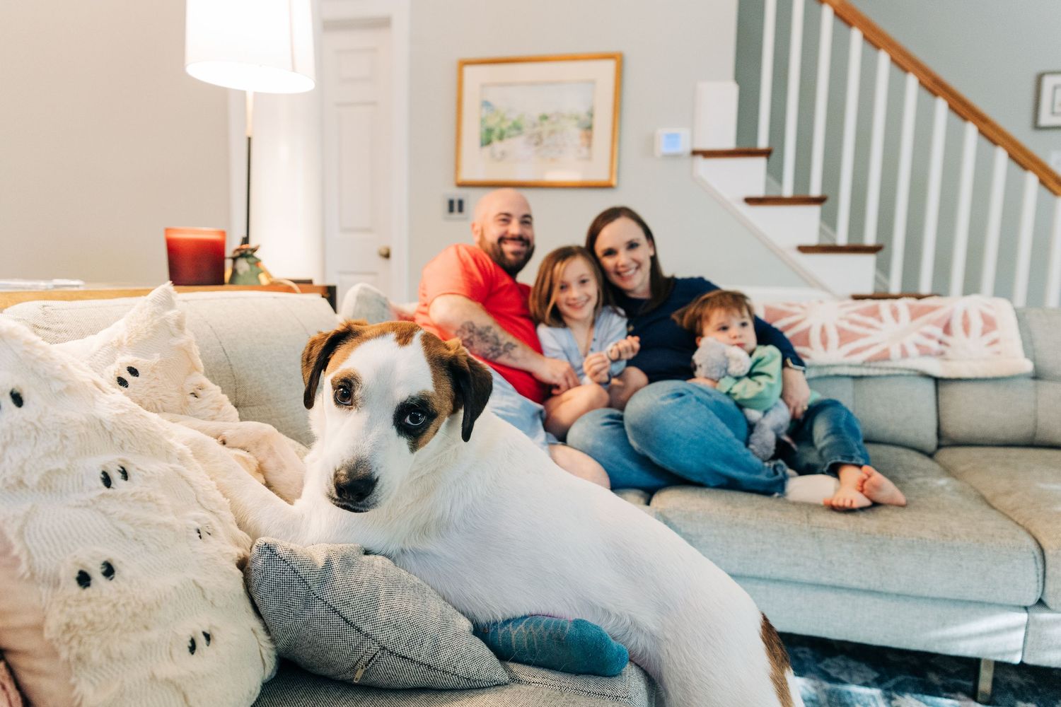 A candid lifestyle photo showing a family relaxing together on a large gray couch in a bright room with a dog posing in front of the image.