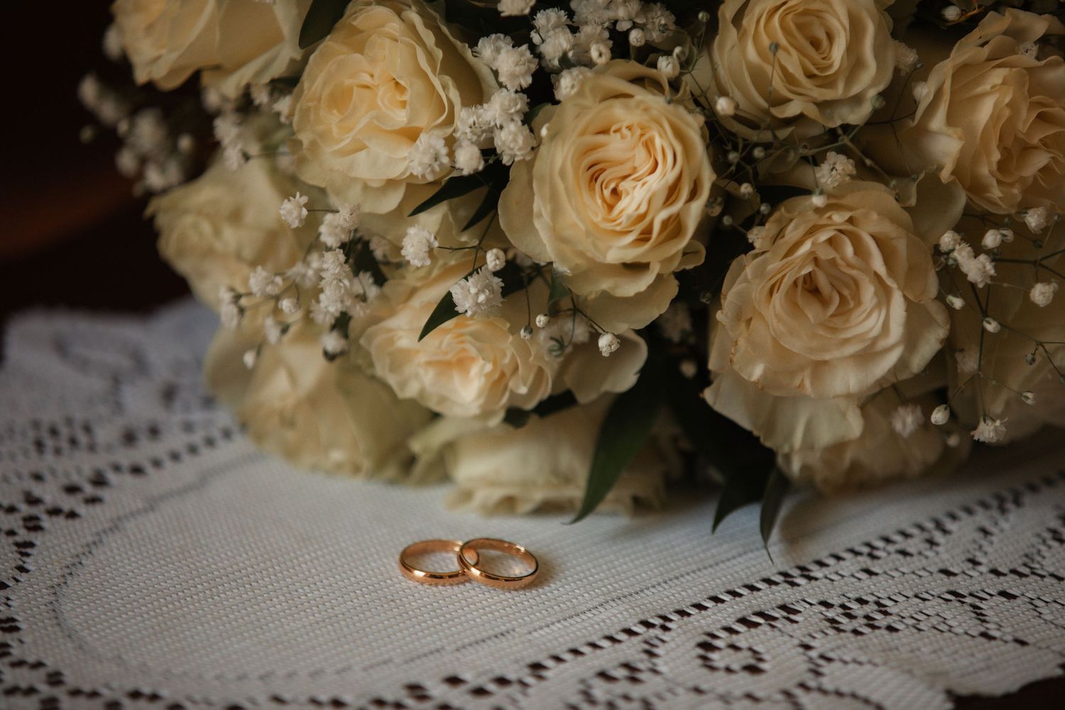 Wedding rings placed on white lace doily with cream roses and baby's breath in background.