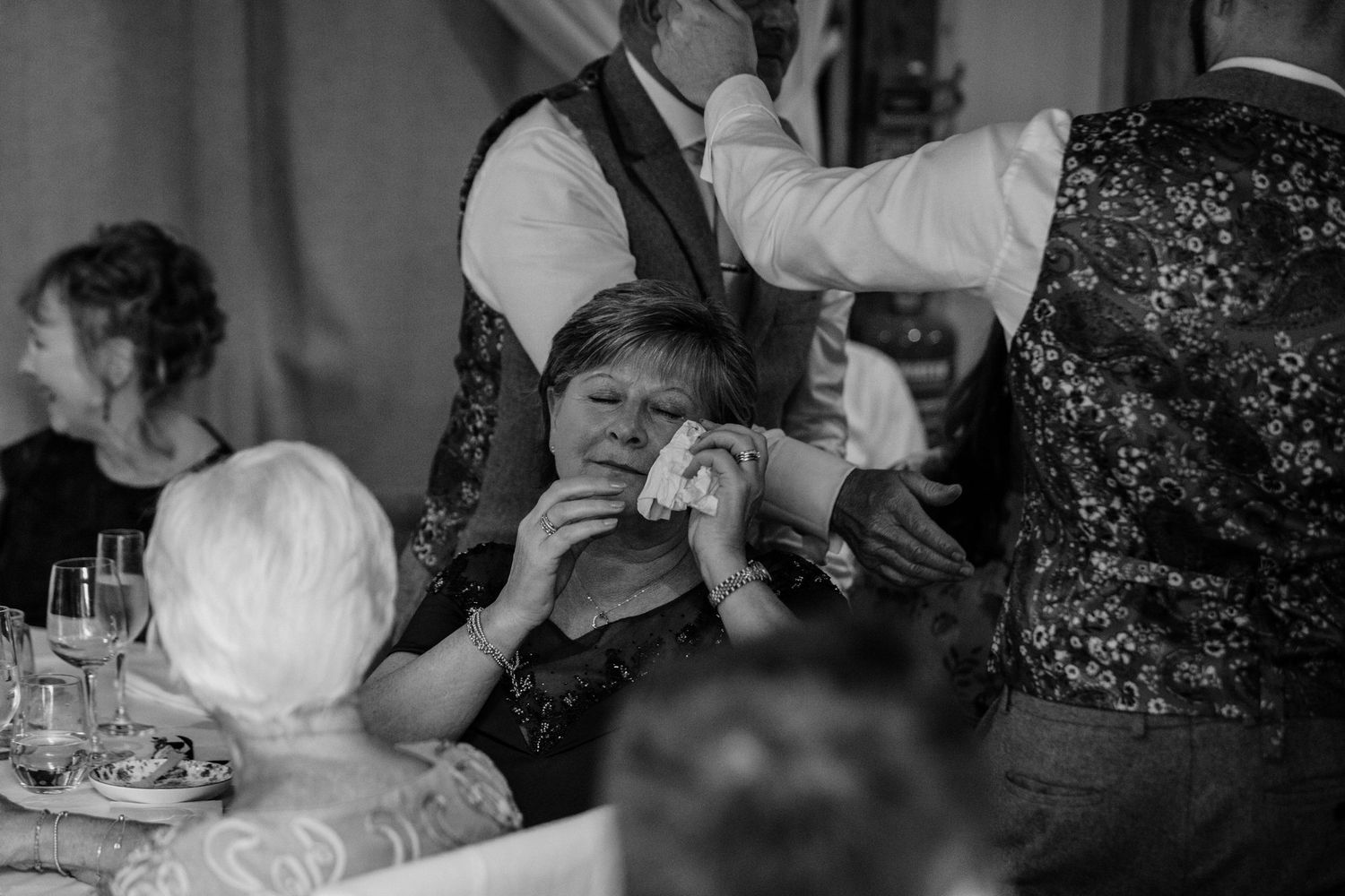 A candid black and white photograph of guests at a wedding reception table showing emotional reactions during speeches.