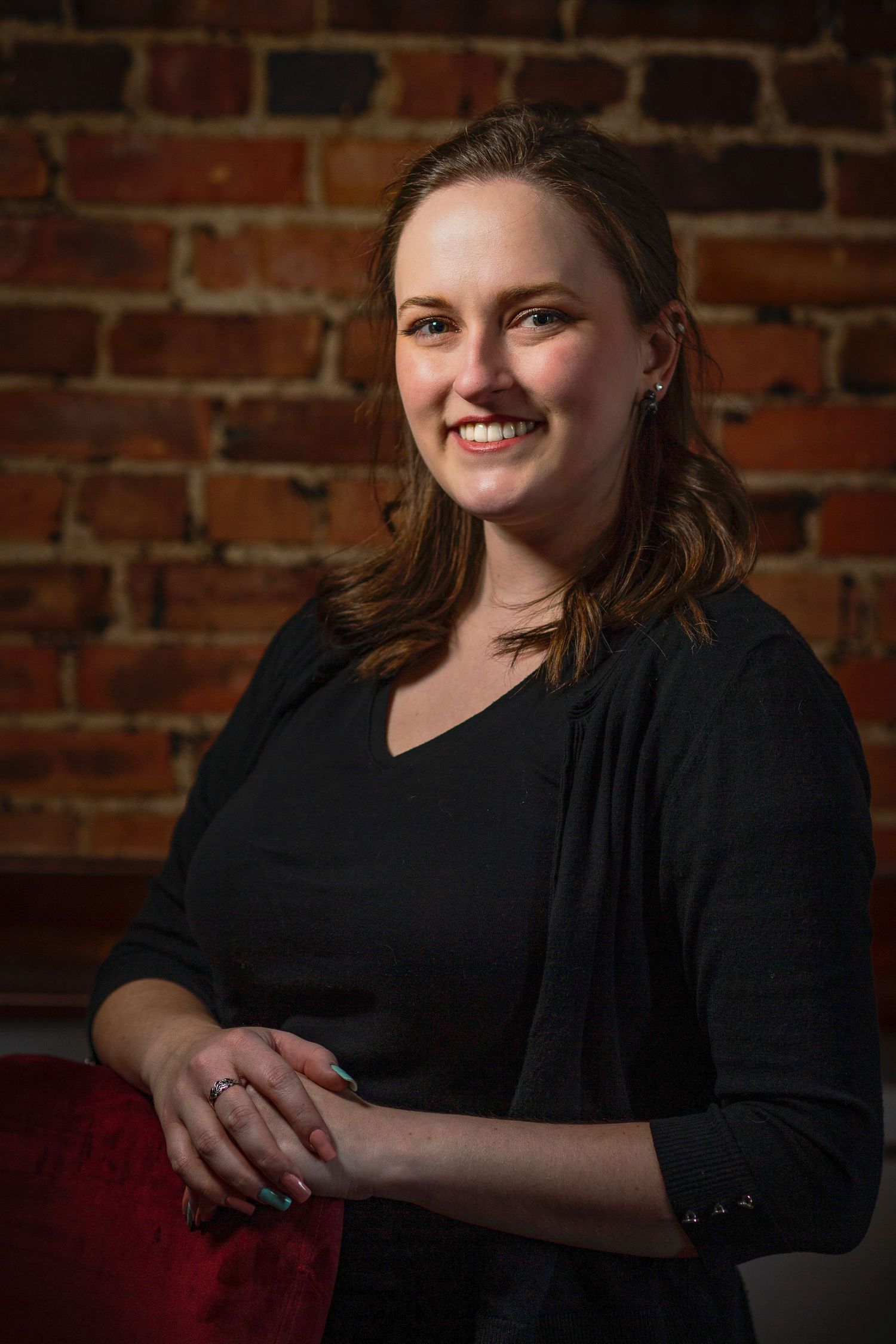 Smiling young woman in a black cardigan posing in front of an exposed red brick wall indoors.