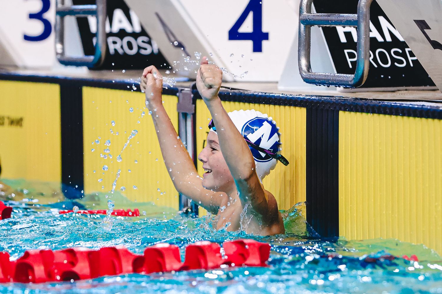 Swimmers racing in a competitive swimming pool with lane dividers and yellow walls in the background.