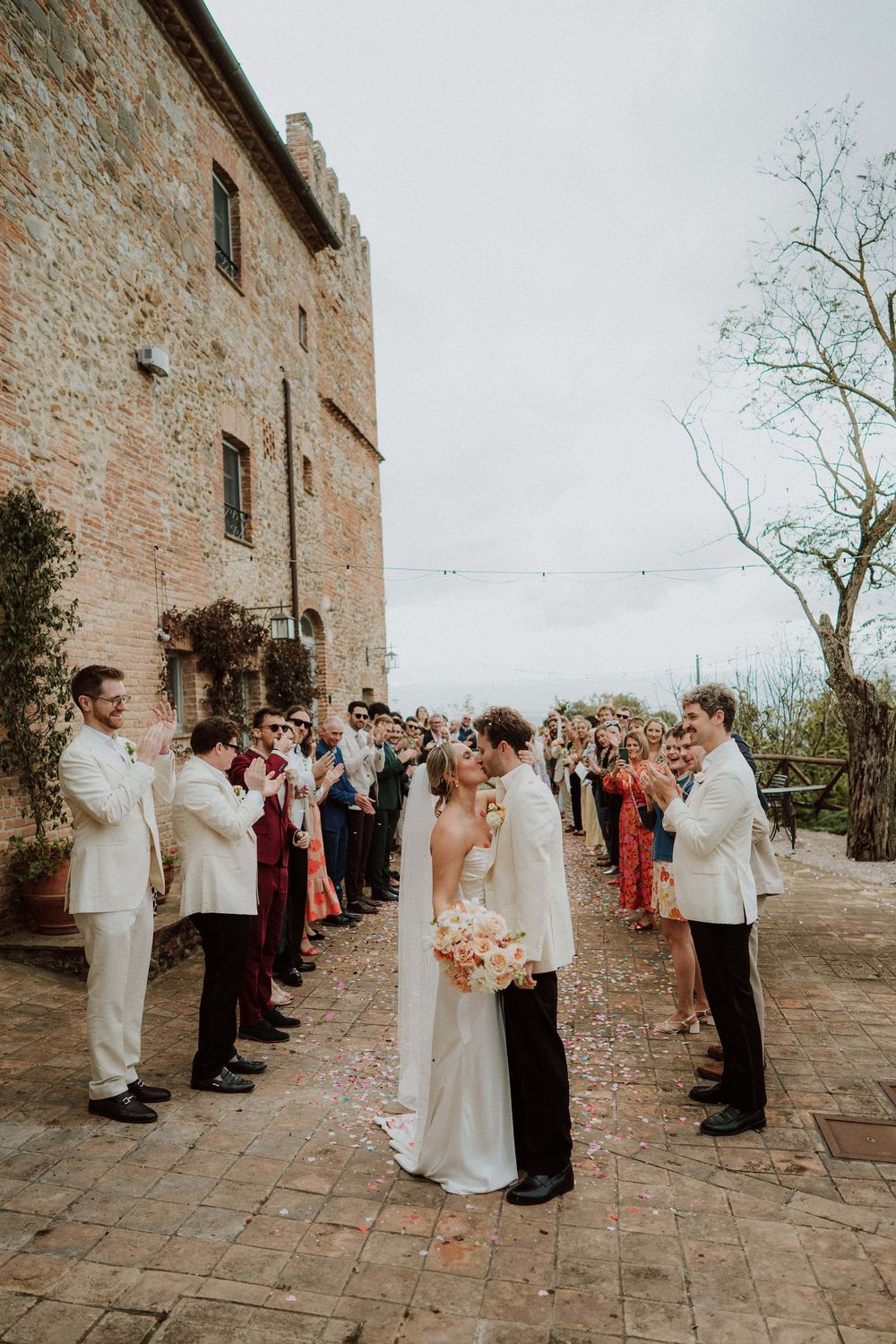 Wedding guests line a stone courtyard to celebrate as a couple shares their first kiss at a rustic Italian villa.