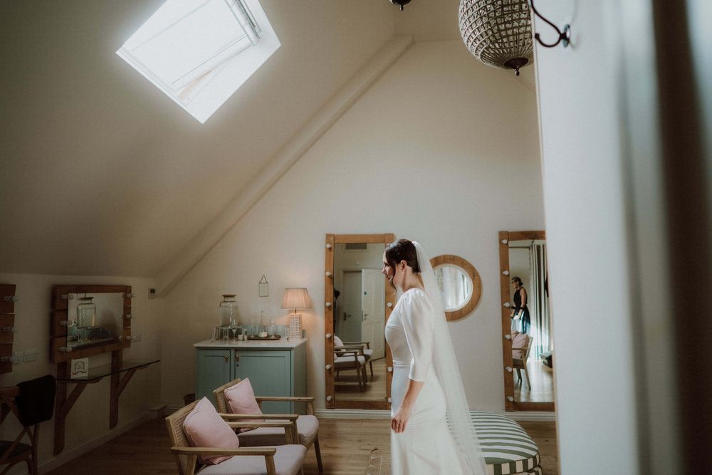 A bride in a white wedding dress stands in a sunlit room preparing for her special day.