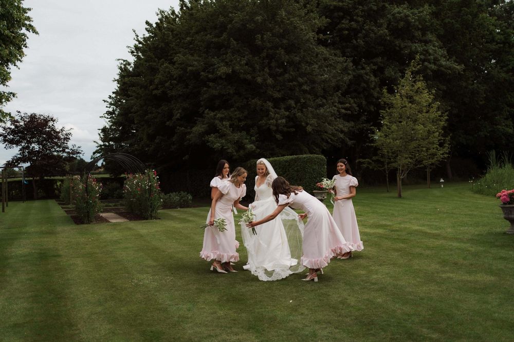 Wedding party members in pink dresses walk together across a lush green lawn at an outdoor celebration.