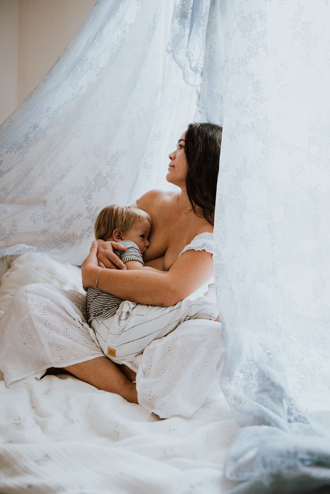 A tender moment captured between parent and child lying together under soft white linen and tulle fabric.