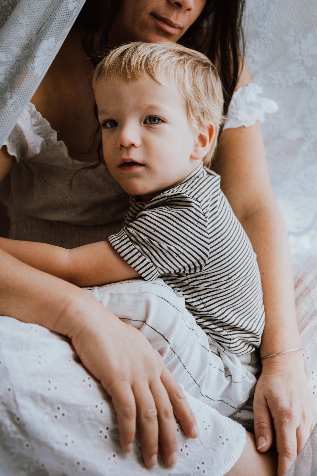 A young child in a striped shirt rests against someone wearing a white dress in a tender indoor portrait.