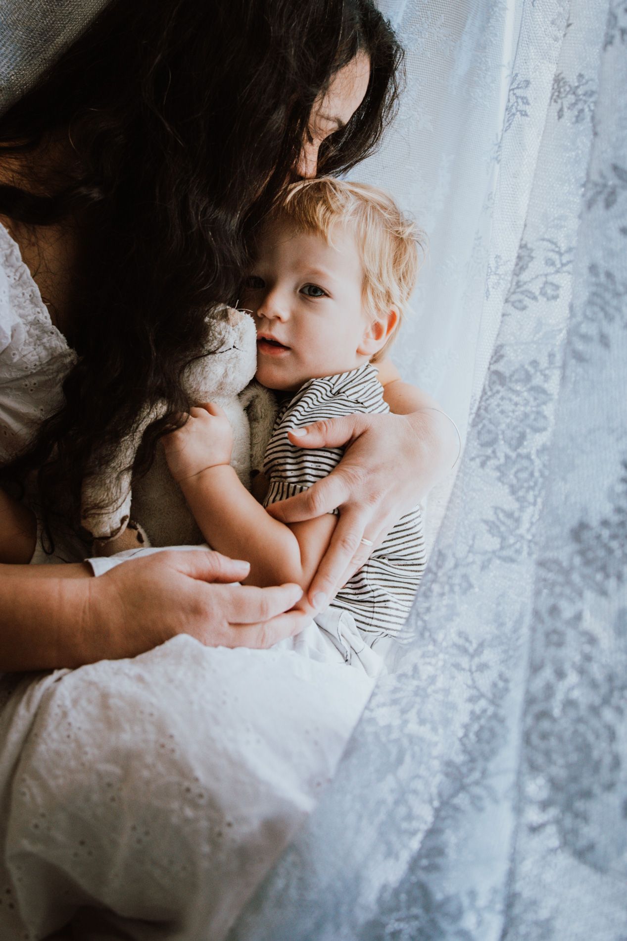 A tender moment captured by a window with lace curtains as parent and child embrace in soft natural light.