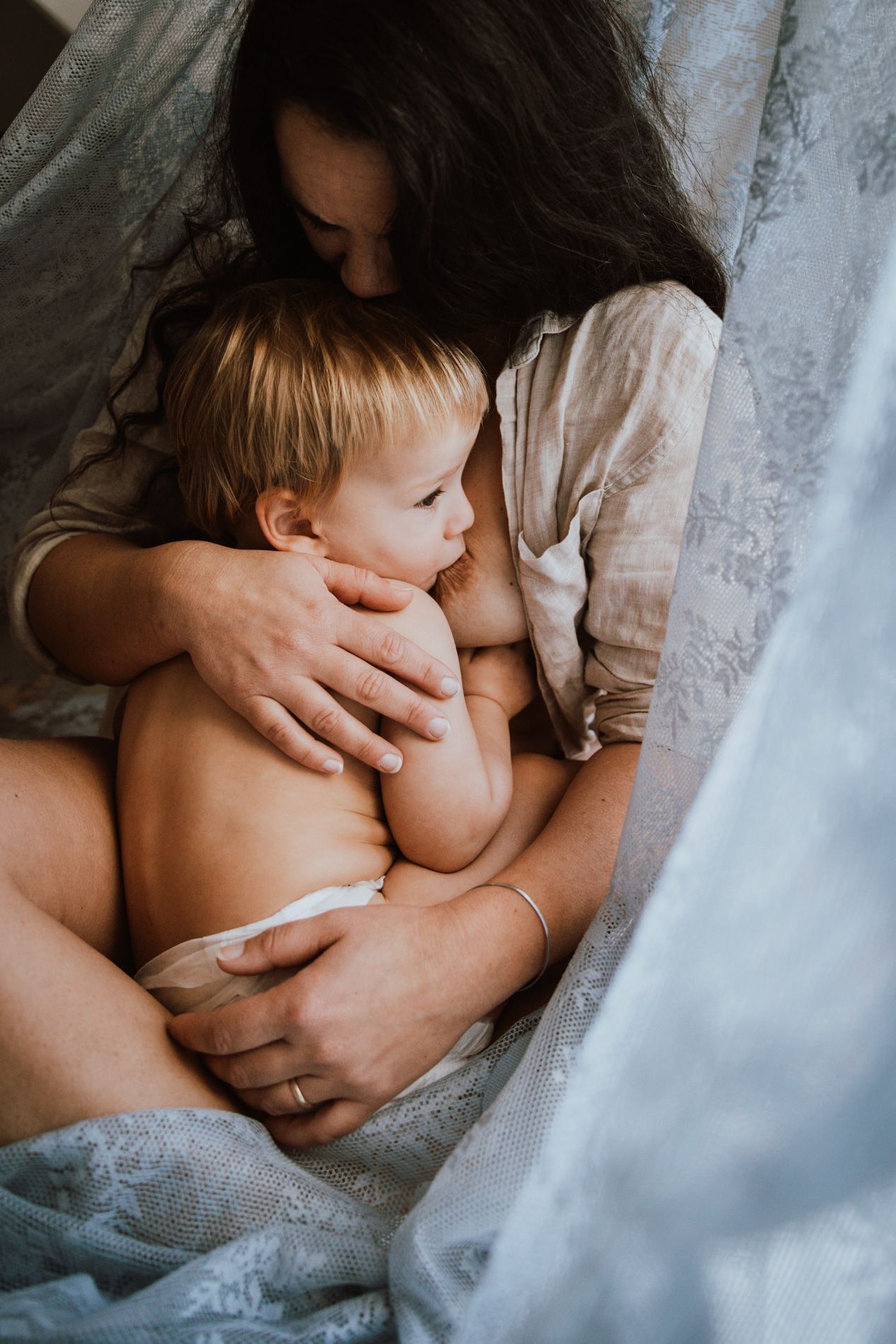 Intimate breastfeeding moment captured in soft natural light with white lace curtain in background.