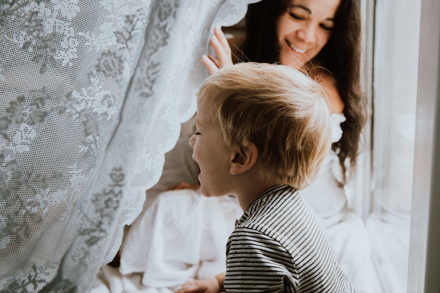 A child in a striped shirt looks up with joy by a window with lace curtains.