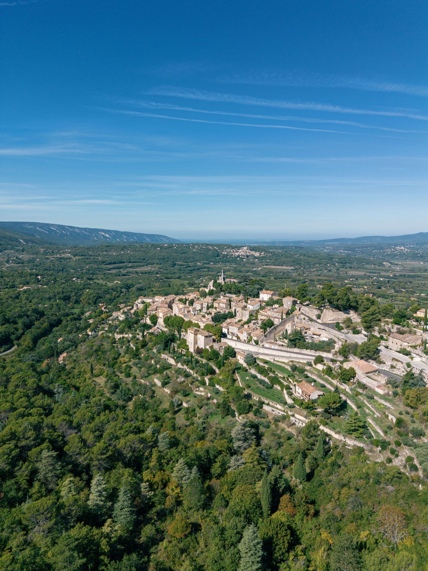 Panoramic aerial view of a hilltop village in Tuscany surrounded by lush green forests under bright blue summer skies.