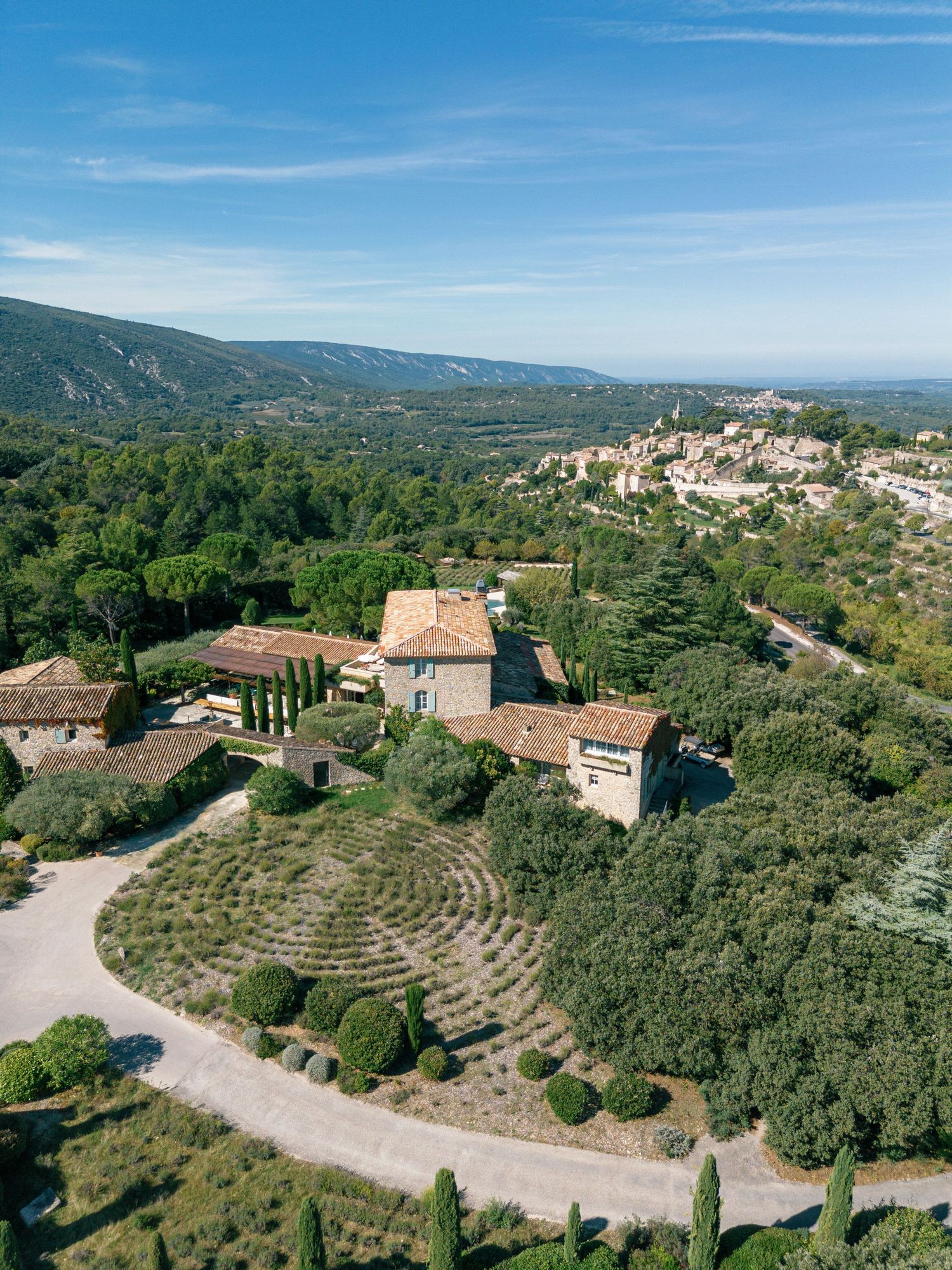 Panoramic aerial view of a hillside village in Provence with terrace gardens and winding roads against mountain backdrop.
