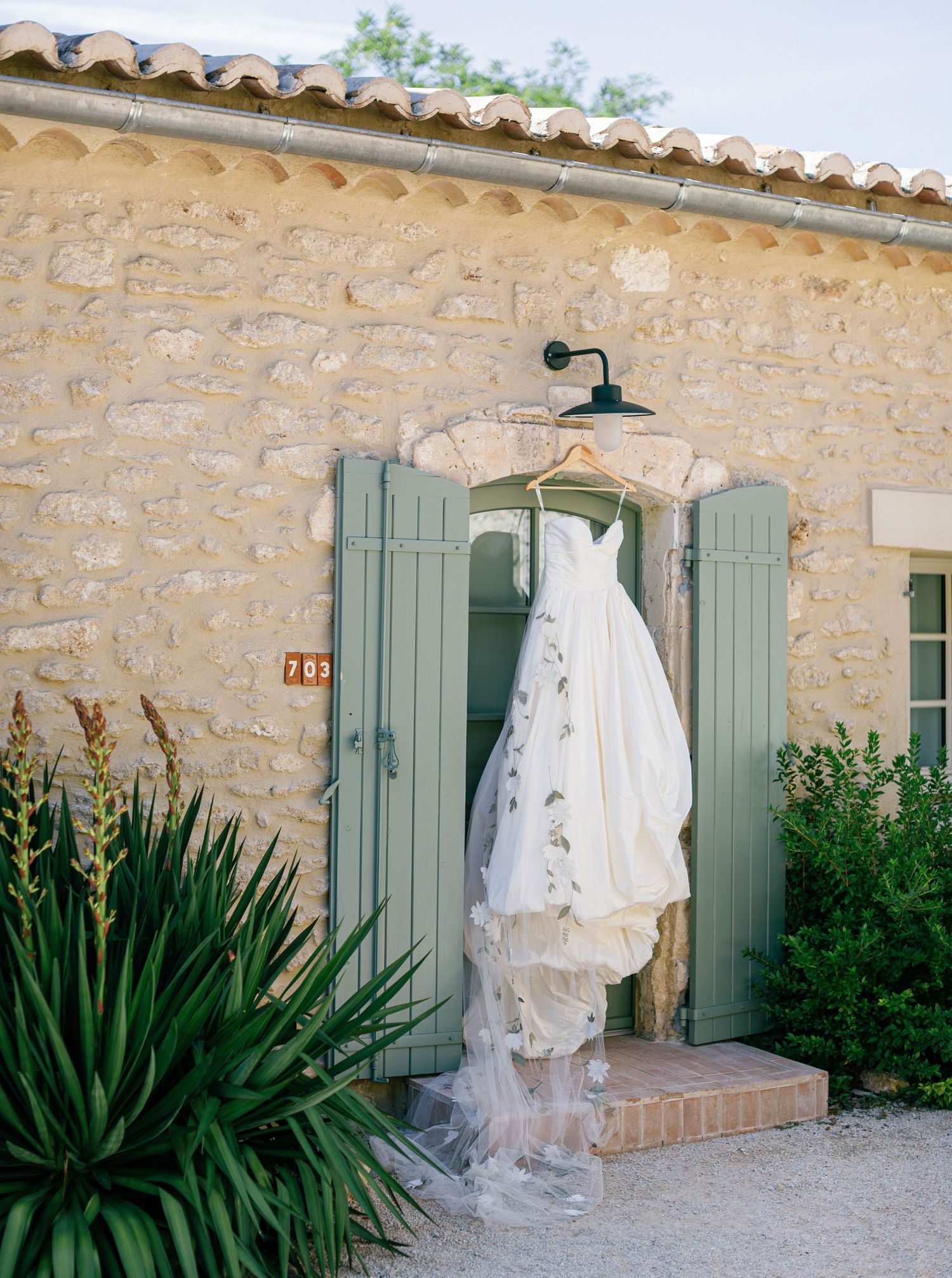 A white wedding dress hangs on a rustic stone wall with green shutters and tropical plants in the foreground.