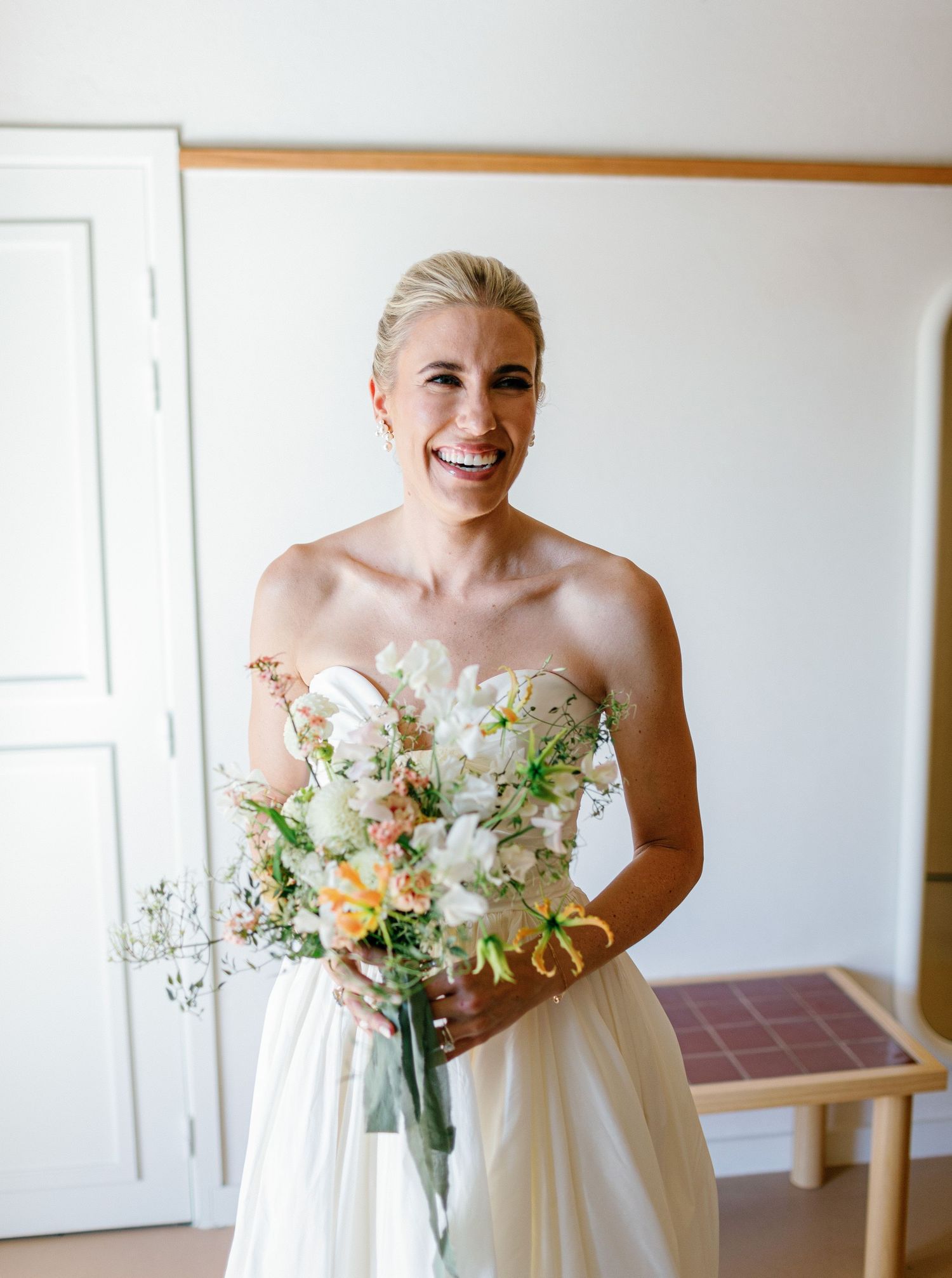 A joyful bride in a strapless white gown holds a delicate bouquet of white and peach flowers against a light wall.