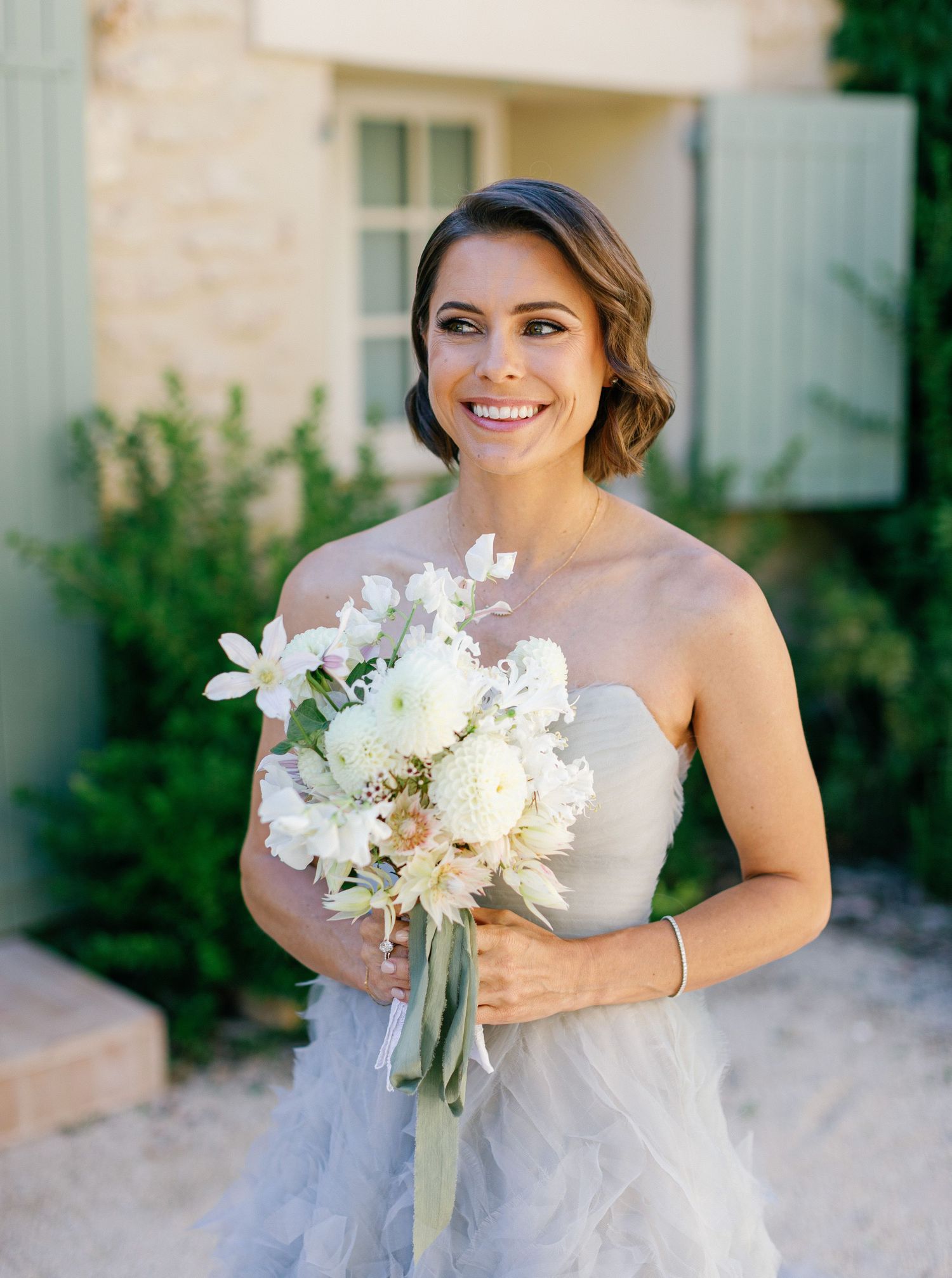 Elegant bride in strapless tulle gown holds a romantic white and blush bouquet with orchids and peonies outdoors.