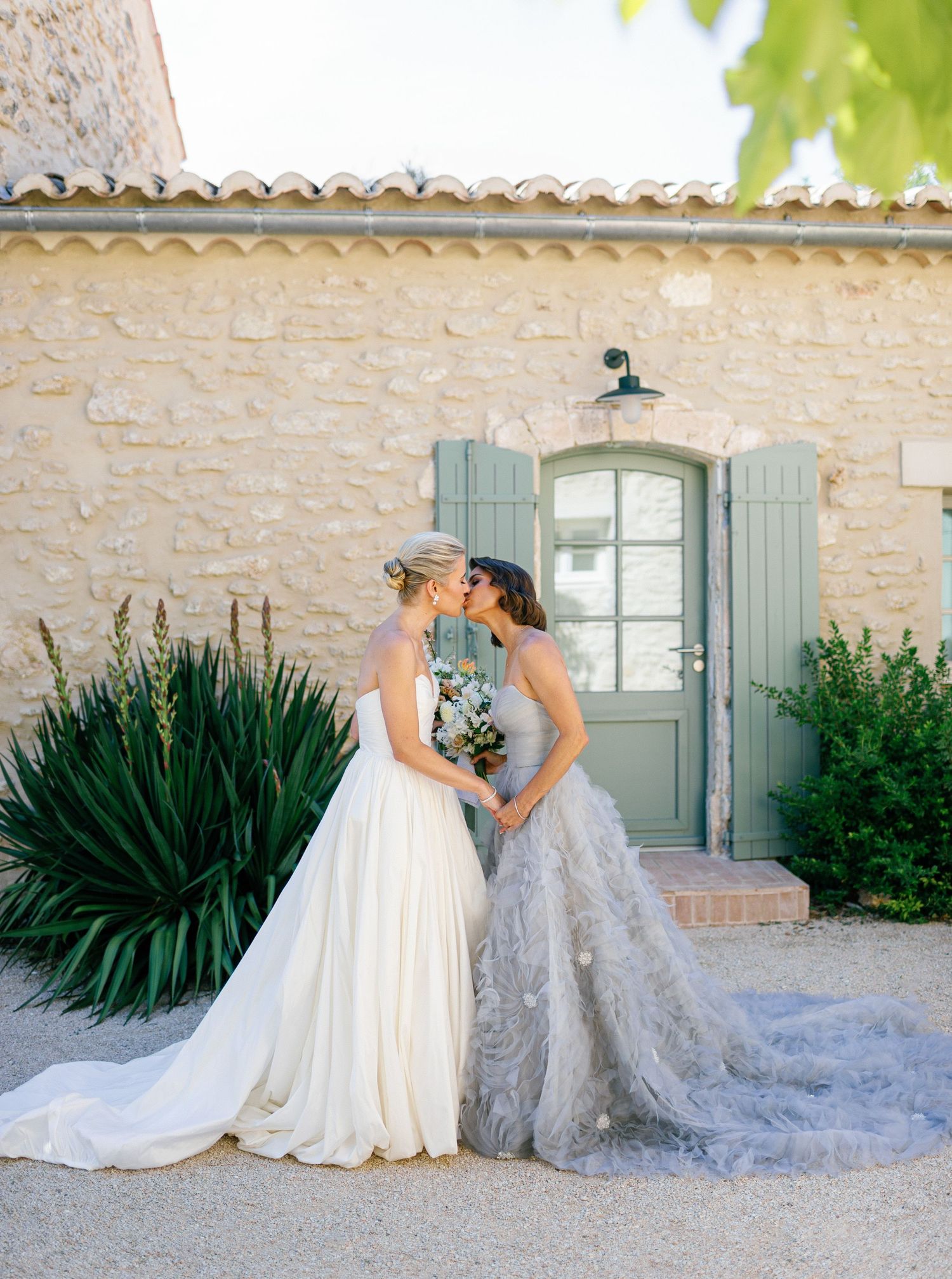 Two elegant wedding gowns with flowing trains create a romantic scene against a rustic stone wall and garden backdrop.