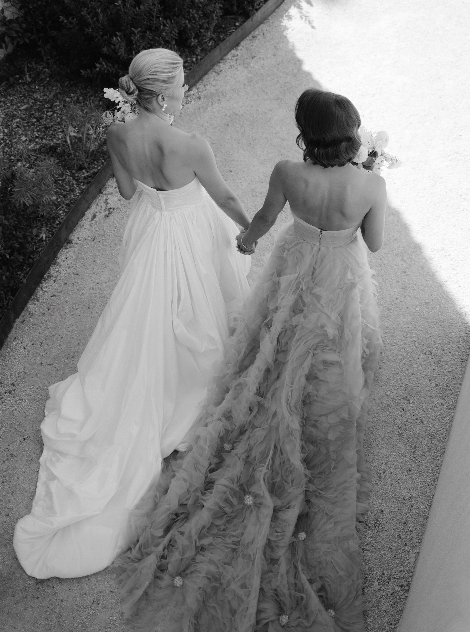 Two people in formal attire walk together outdoors, one wearing a flowing white gown and the other in elegant attire.