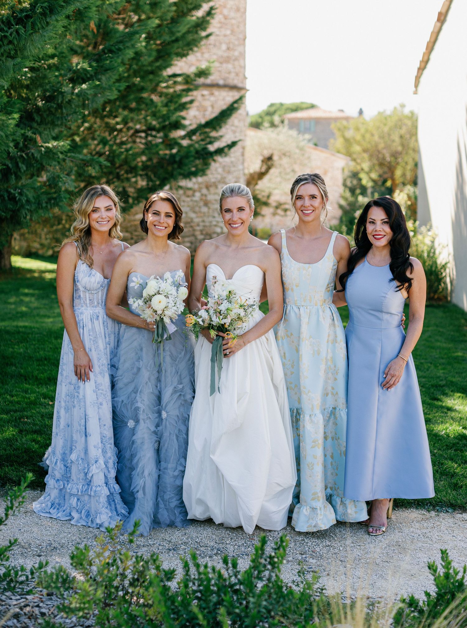 Bridesmaids in soft blue floral dresses pose with the bride in a white gown during an outdoor summer wedding.
