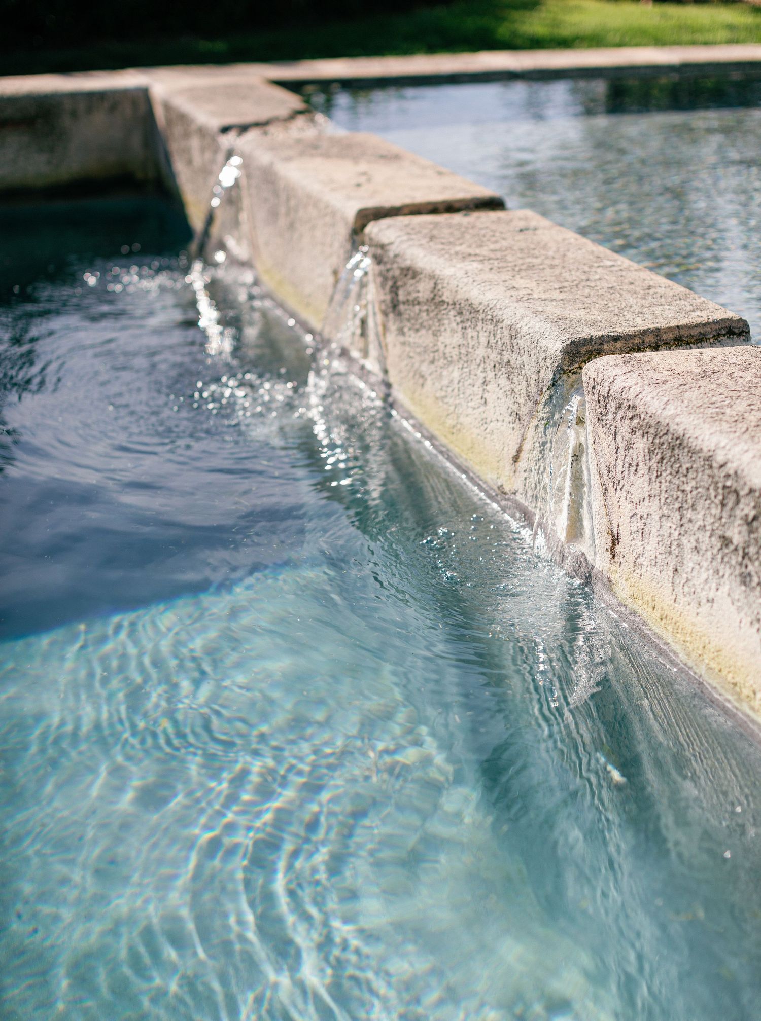 Water cascades over a concrete ledge into a pool creating a serene waterfall effect in bright sunlight.