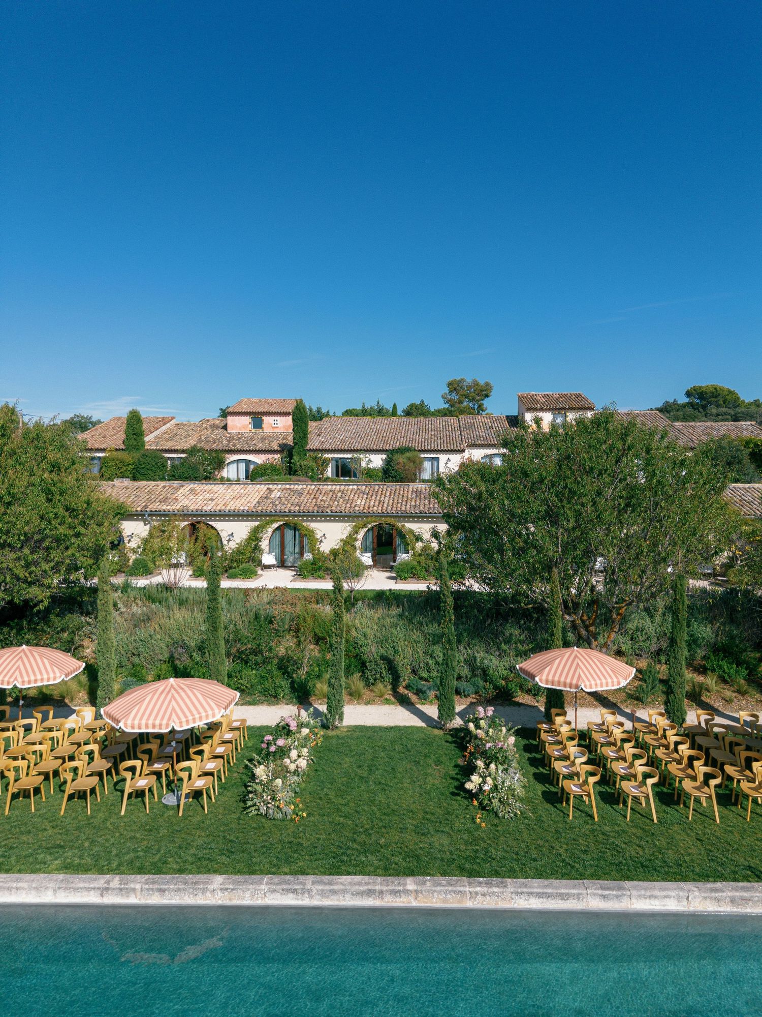 Panoramic view of an outdoor wedding ceremony setup with wooden chairs arranged on a lawn against a Mediterranean villa backdrop.