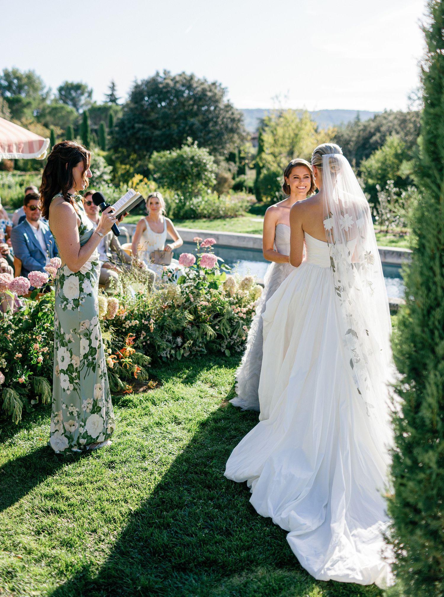 Wedding party walking along a scenic garden path with floral arrangements and greenery on a sunny day.