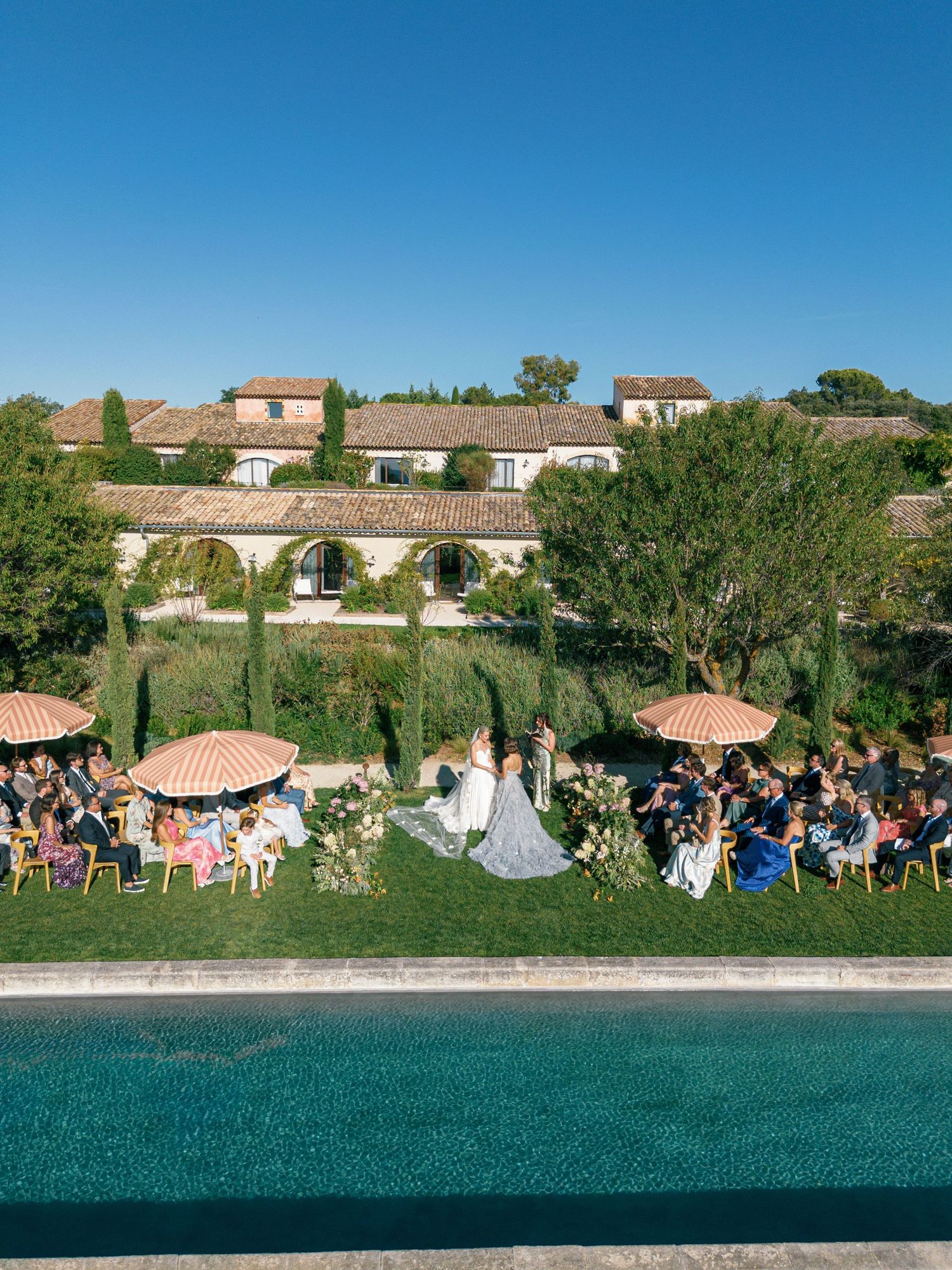 An outdoor wedding ceremony by a pool with guests seated under orange umbrellas at a Mediterranean-style venue.