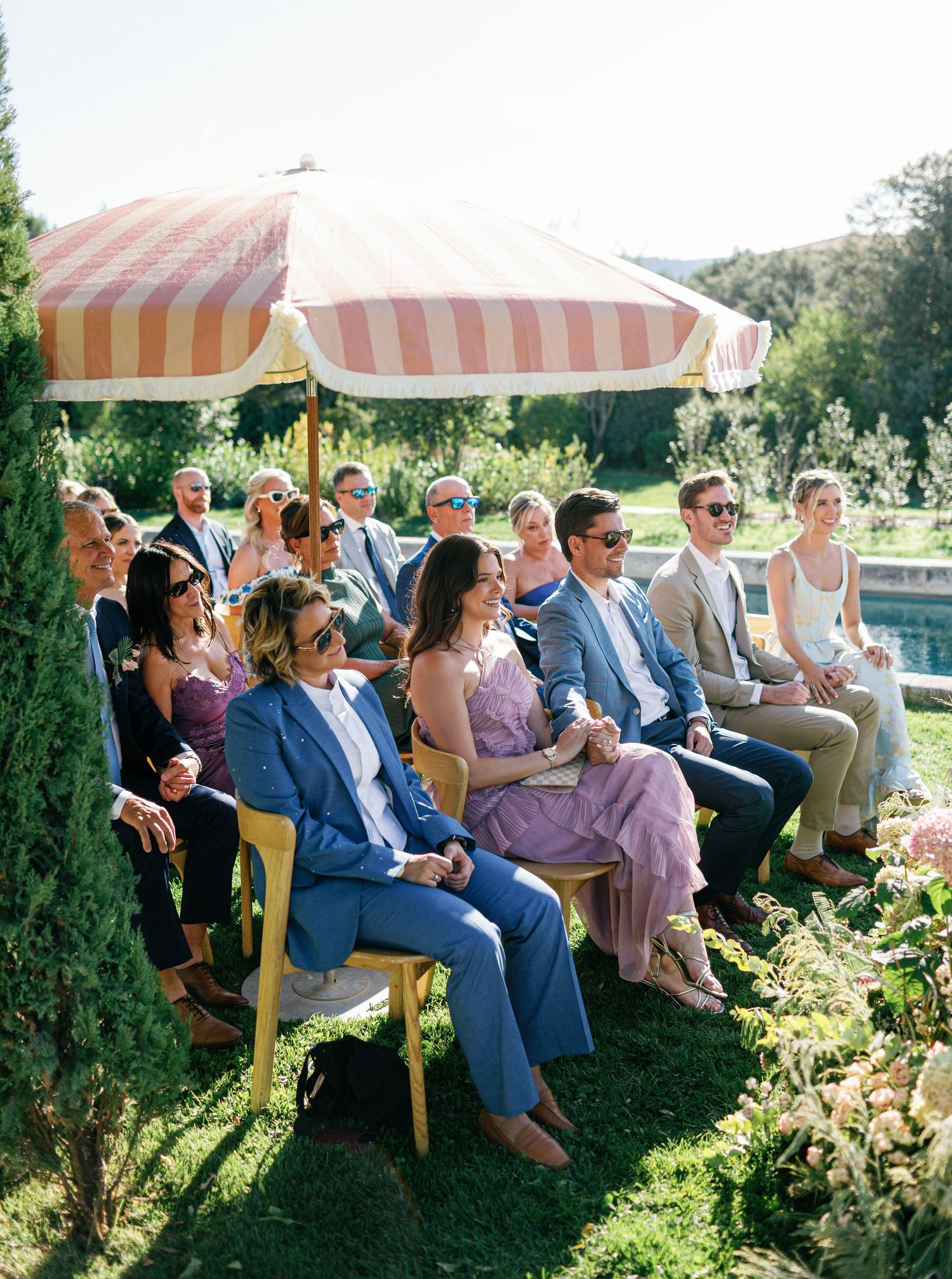Wedding guests seated outdoors under striped umbrellas in a garden setting during a summer ceremony.