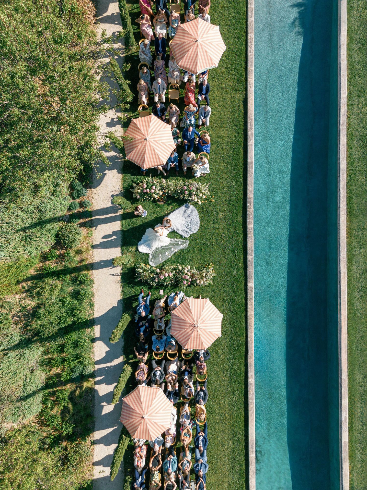 Aerial view of people gathered under pink umbrellas along a turquoise pool on a sunny day.