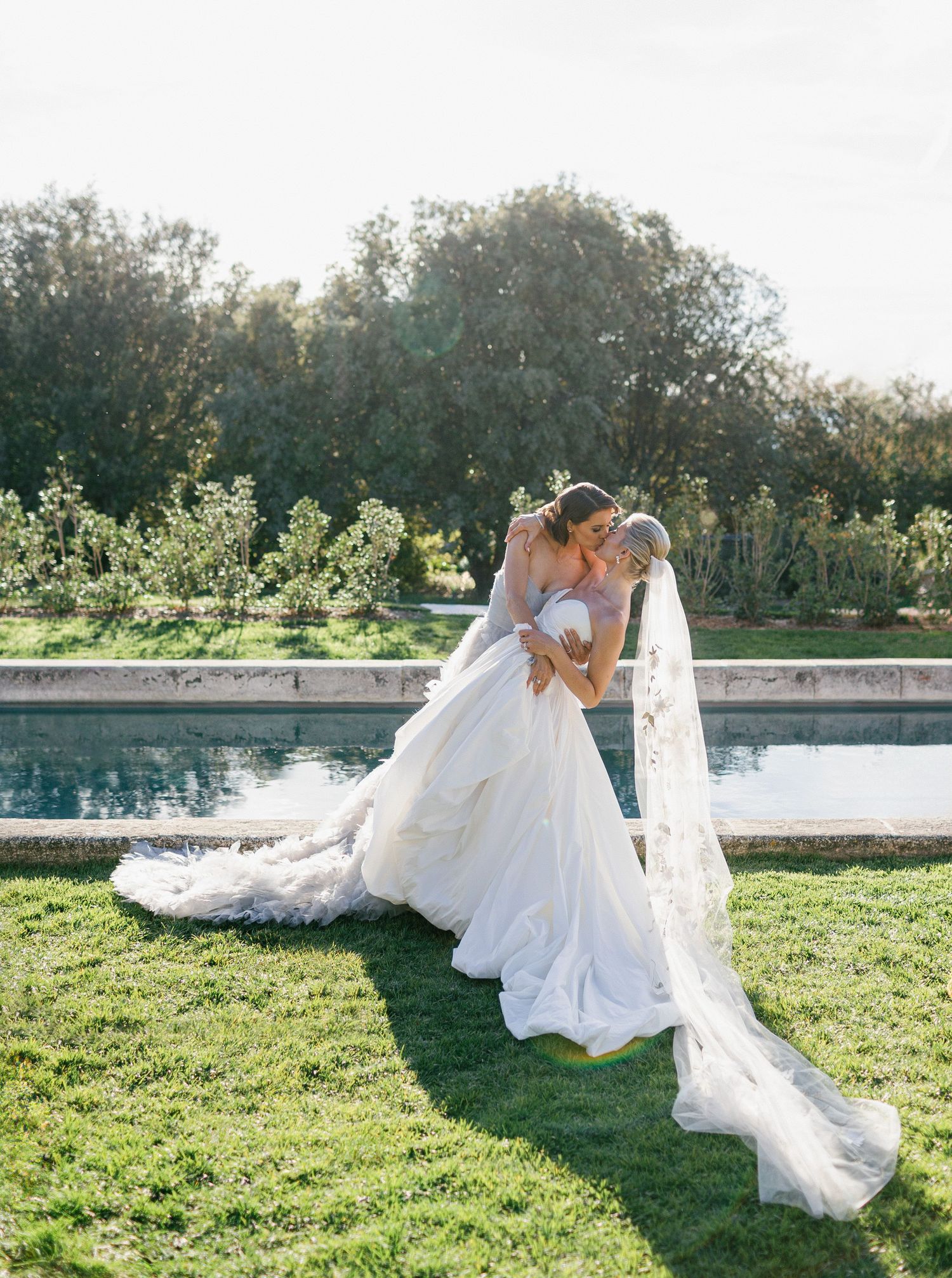 Flowing white wedding dress and veil captured in motion against a natural outdoor backdrop with grass and trees.