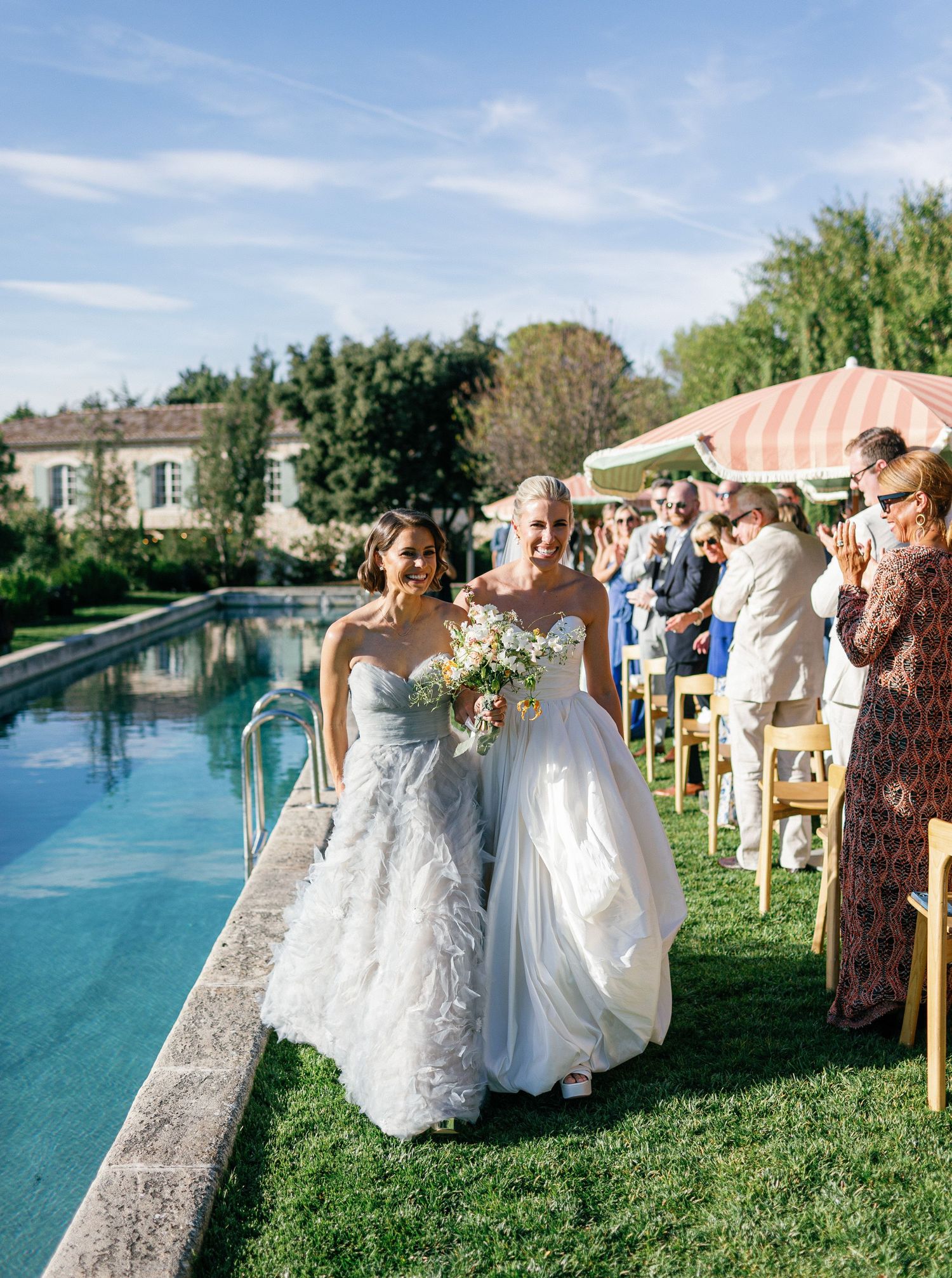 Wedding party poses for photos by a serene pool on a sunny day with green grass and trees in the background.
