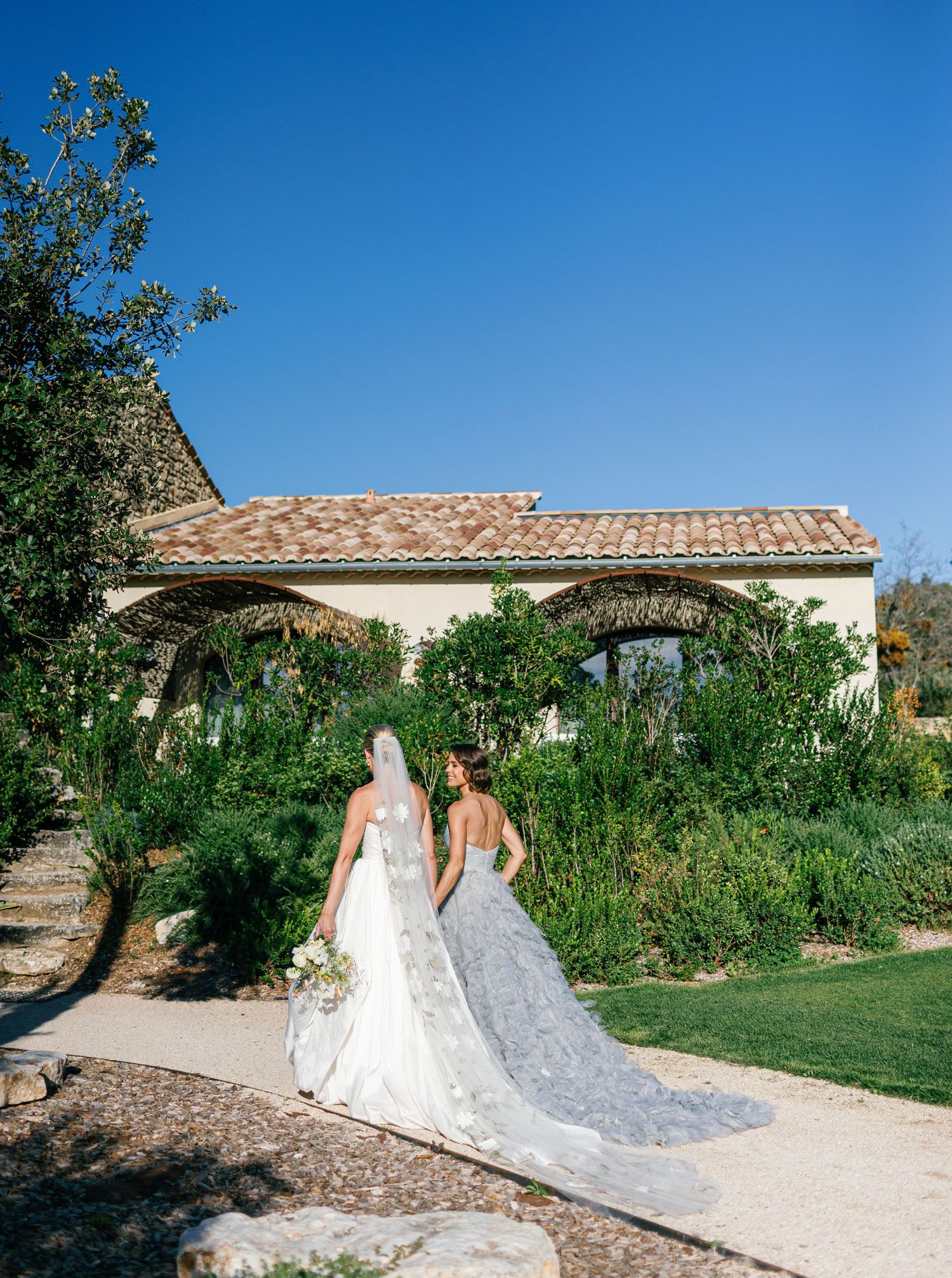 Two elegant white wedding dresses with long trains flow across a stone path in a Mediterranean garden setting.