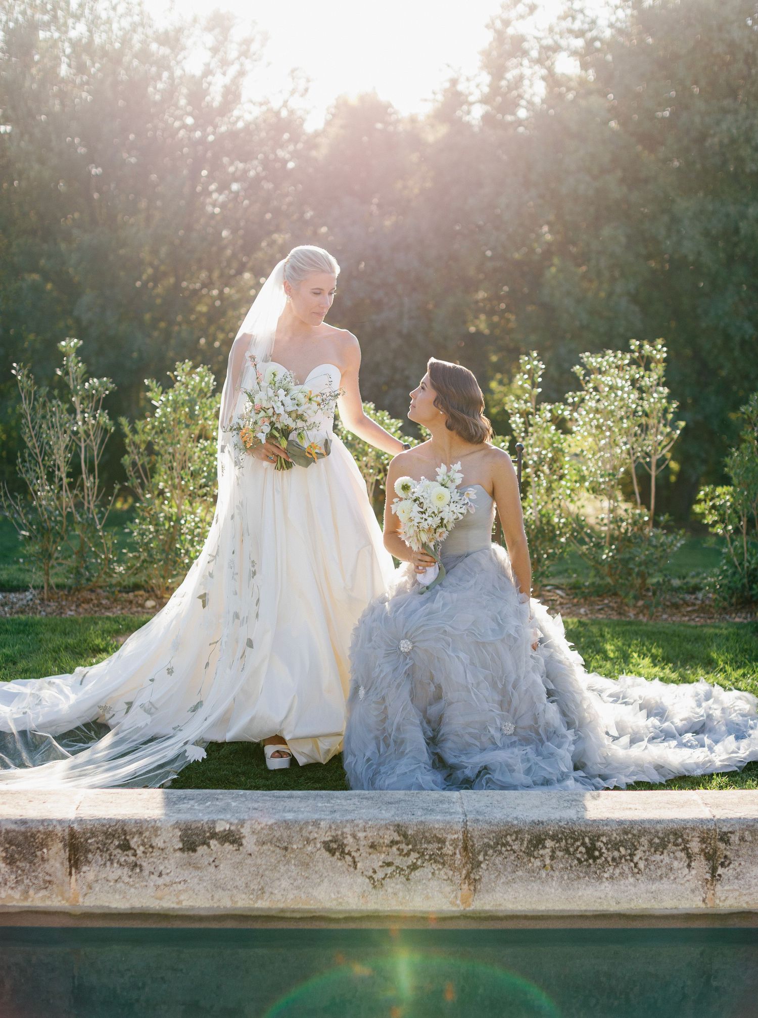 Two brides in elegant white wedding gowns with flowing trains sit together by a pool during golden hour sunset.