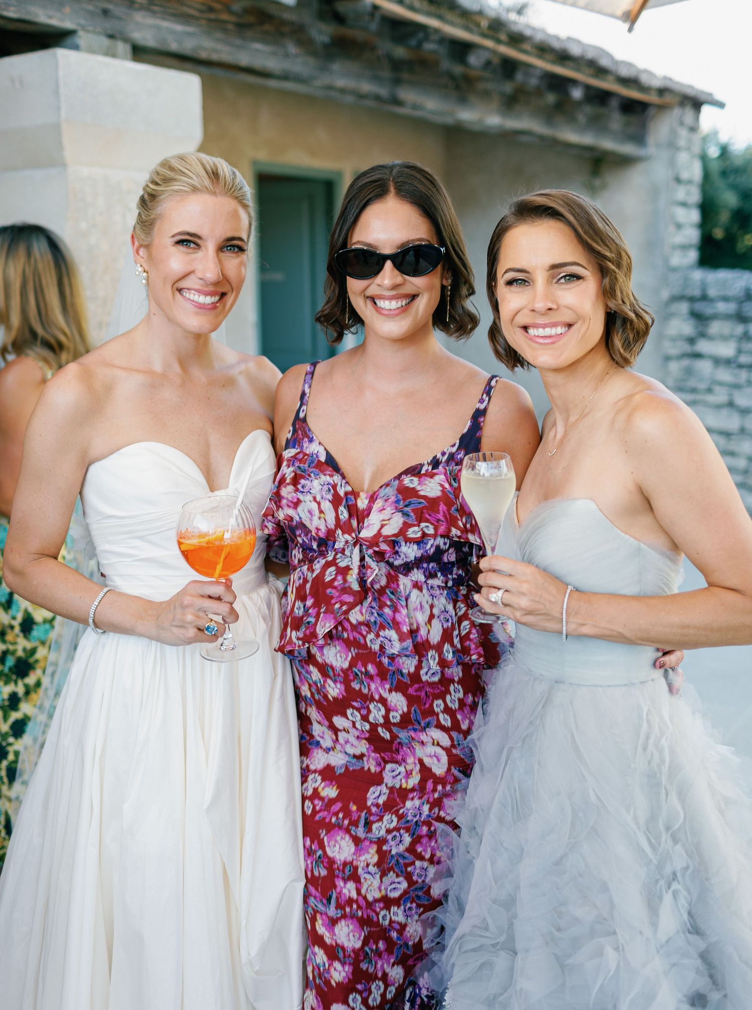 Three women in formal attire hold cocktails while celebrating at an outdoor wedding reception.