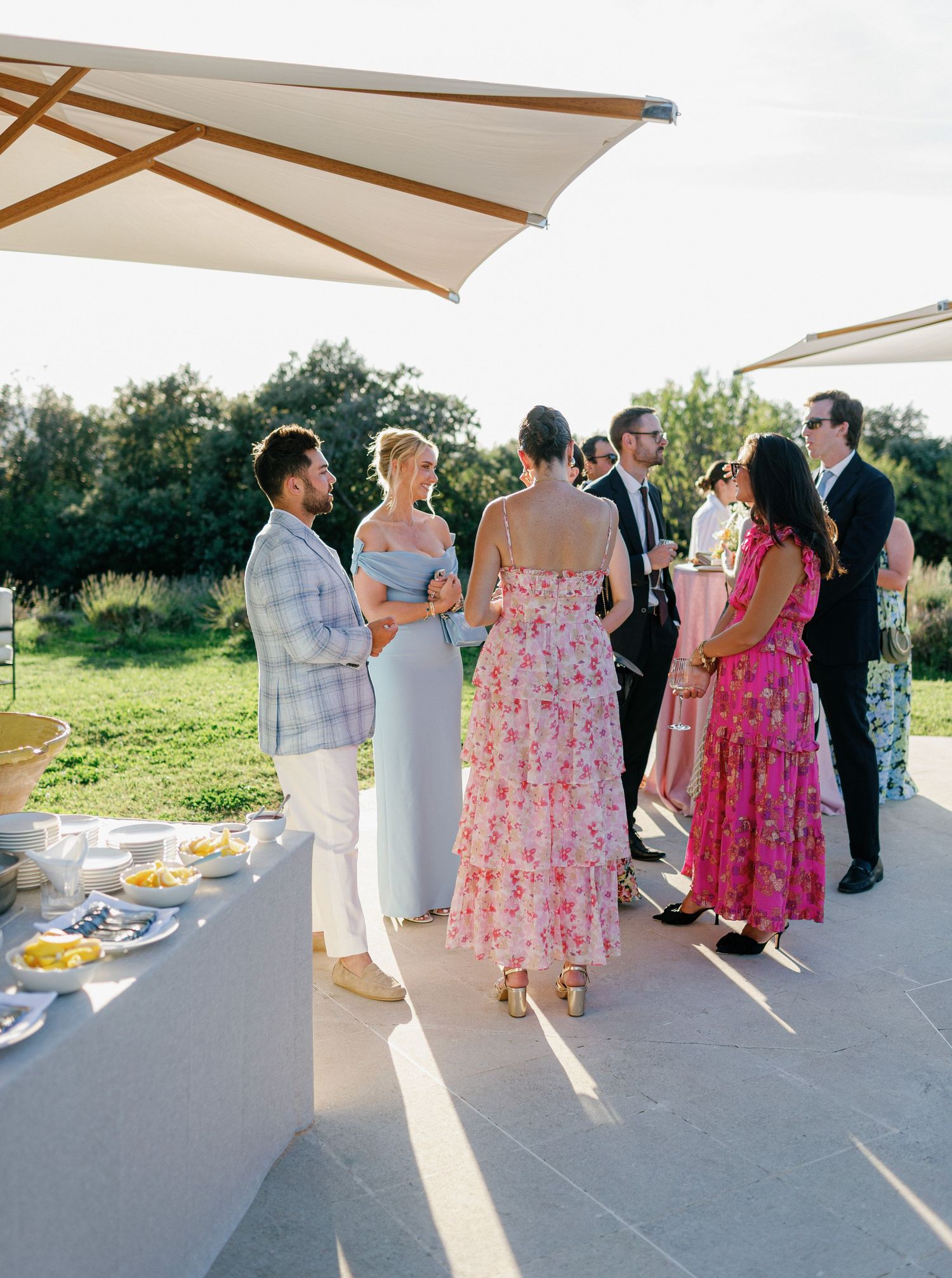 A line of guests at an outdoor reception buffet stand under white umbrellas on a sunny afternoon.