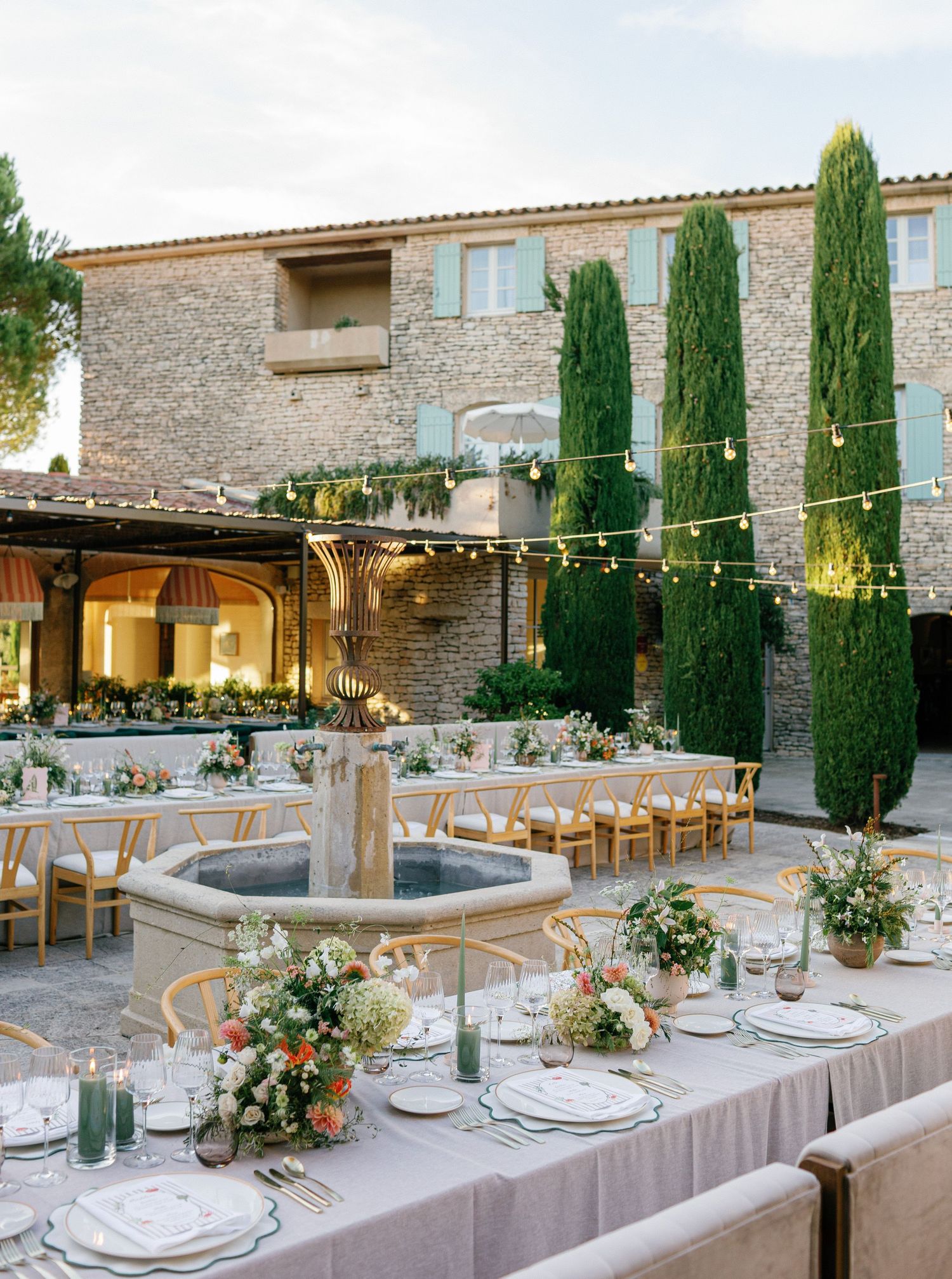Elegant outdoor wedding reception setup with stone building backdrop, cypress trees and string lights over dining tables.