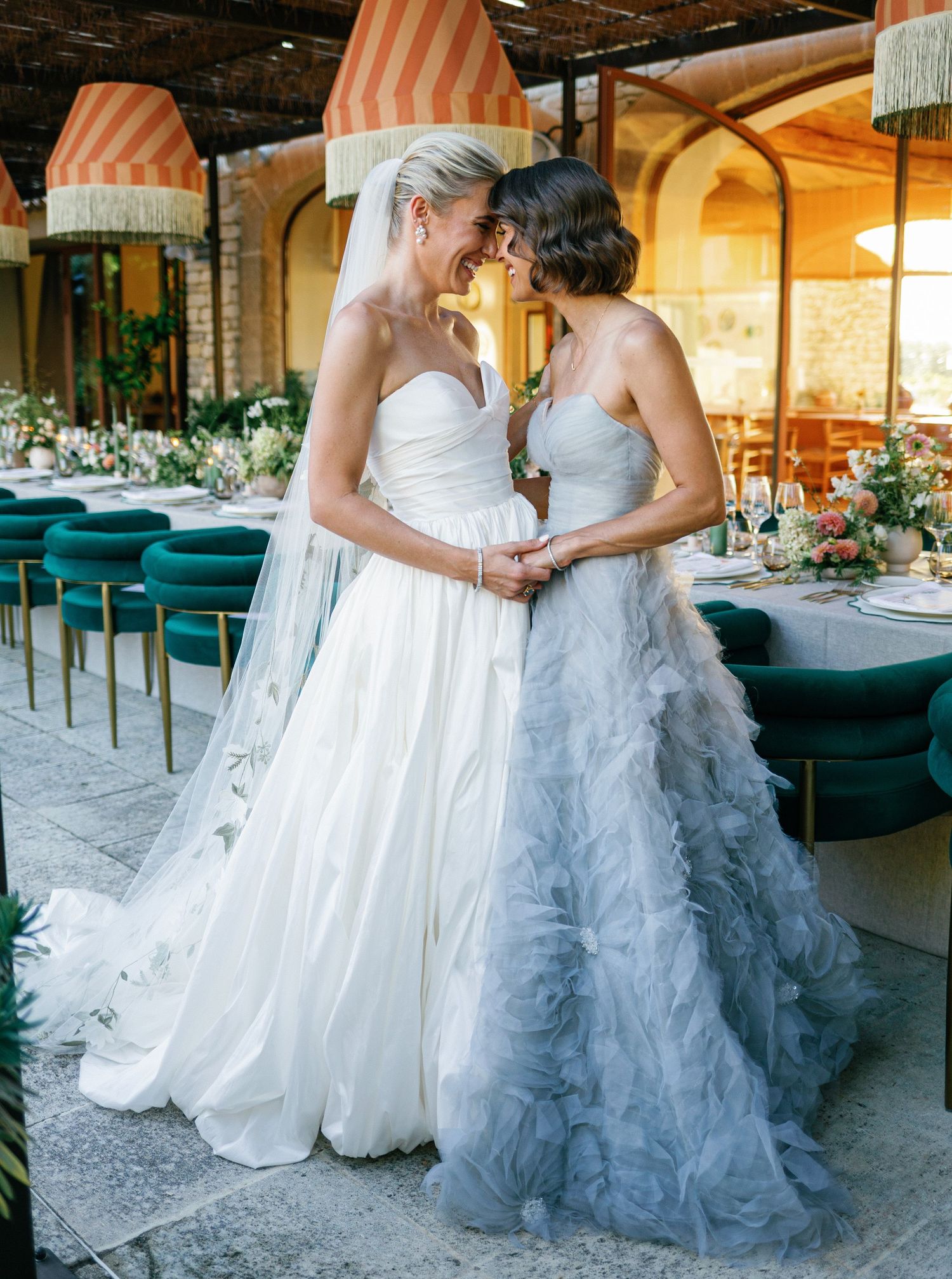 Two brides in white and gray ruffled wedding dresses hold hands during an elegant indoor reception.