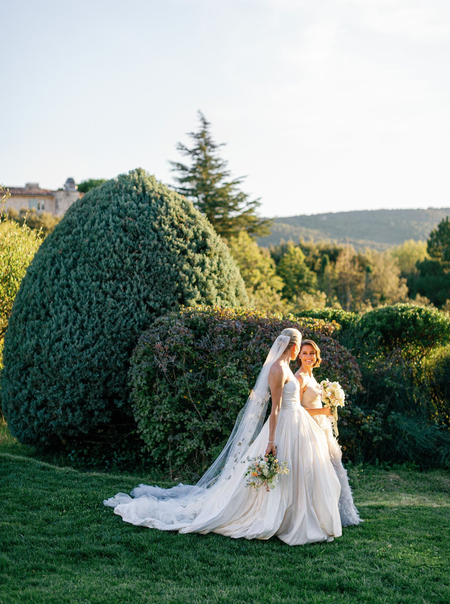A couple in wedding attire poses together on a grassy hillside with manicured hedges and trees at sunset.