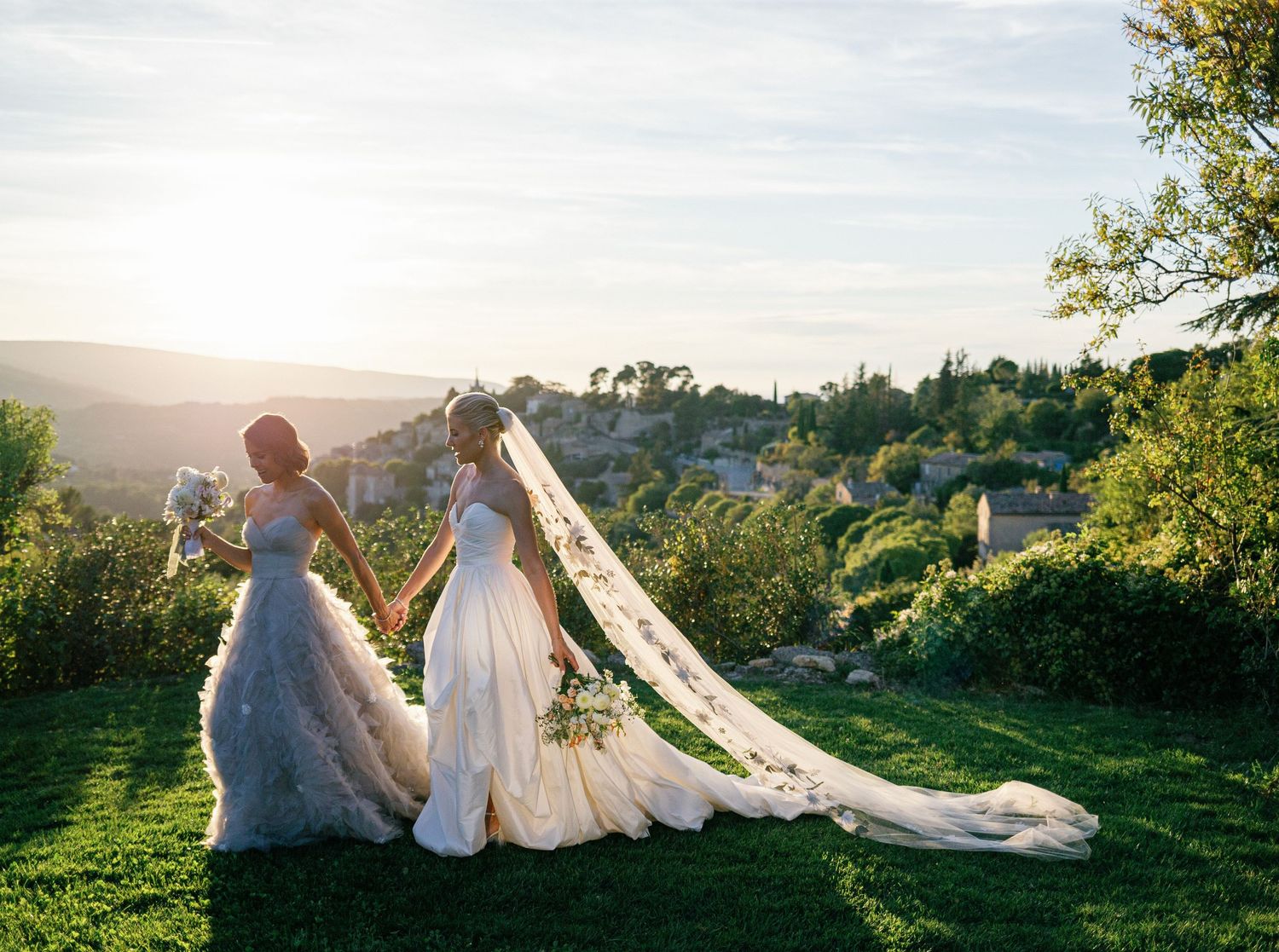 A couple in wedding attire stands together in a scenic hilltop garden during golden hour sunset.